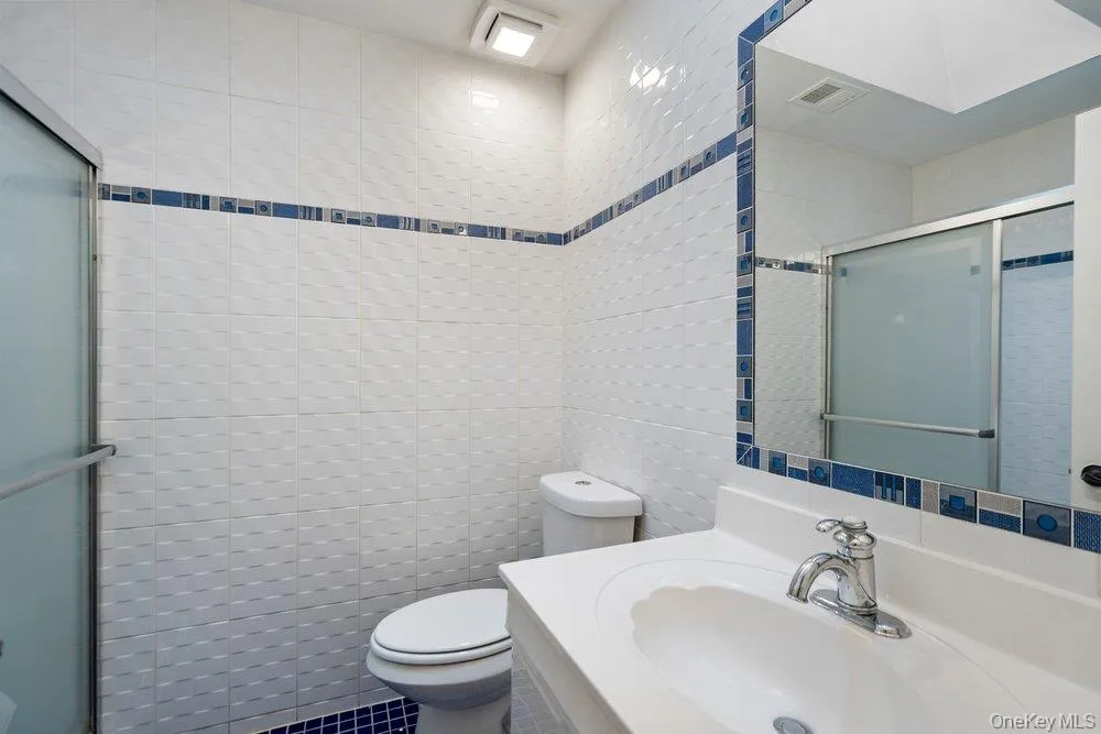 Bathroom featuring white textured wall tiles with a blue decorative band, a white vanity with an integrated sink, a chrome faucet, a framed mirror with a matching blue tile accent, and a shower enclosure with frosted glass sliding doors Bathroom featuring white textured wall tiles with a blue decorative band, a white vanity with an integrated sink, a chrome faucet, a framed mirror with a matching blue tile accent, and a shower enclosure with frosted glass sliding doors