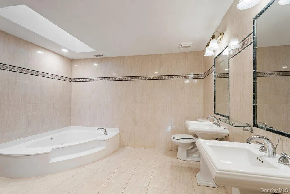 Spacious bathroom featuring a corner jetted tub, dual pedestal sinks, a skylight, and recessed lighting Spacious bathroom featuring a corner jetted tub, dual pedestal sinks, a skylight, and recessed lighting