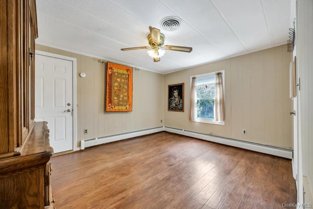 Spacious room featuring wood-finish flooring, light-toned vertical paneling, and a ceiling fan with integrated lighting Spacious room featuring wood-finish flooring, light-toned vertical paneling, and a ceiling fan with integrated lighting