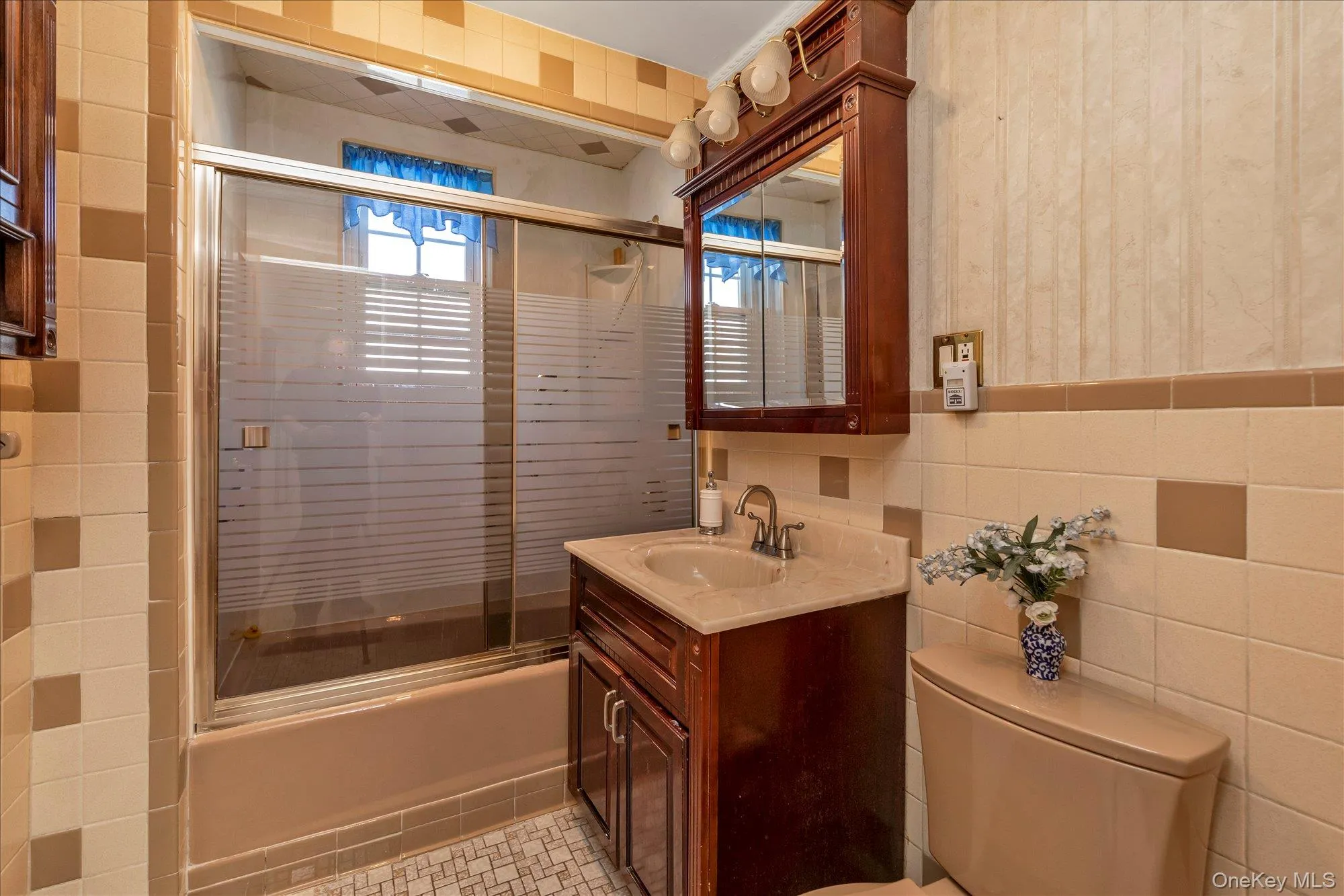 Bathroom featuring a vanity with a light-colored countertop and dark wood-finish cabinetry, a built-in medicine cabinet with integrated lighting, a shower-tub combination with sliding frosted glass doors, and light-toned tile flooring Bathroom featuring a vanity with a light-colored countertop and dark wood-finish cabinetry, a built-in medicine cabinet with integrated lighting, a shower-tub combination with sliding frosted glass doors, and light-toned tile flooring