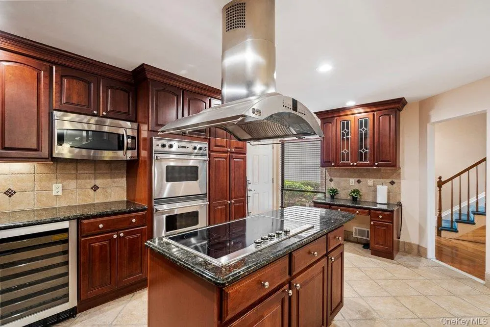 Kitchen featuring a large central island with a built-in cooktop, stainless steel range hood, stainless steel double wall ovens, built-in microwave, and a wine refrigerator Kitchen featuring a large central island with a built-in cooktop, stainless steel range hood, stainless steel double wall ovens, built-in microwave, and a wine refrigerator