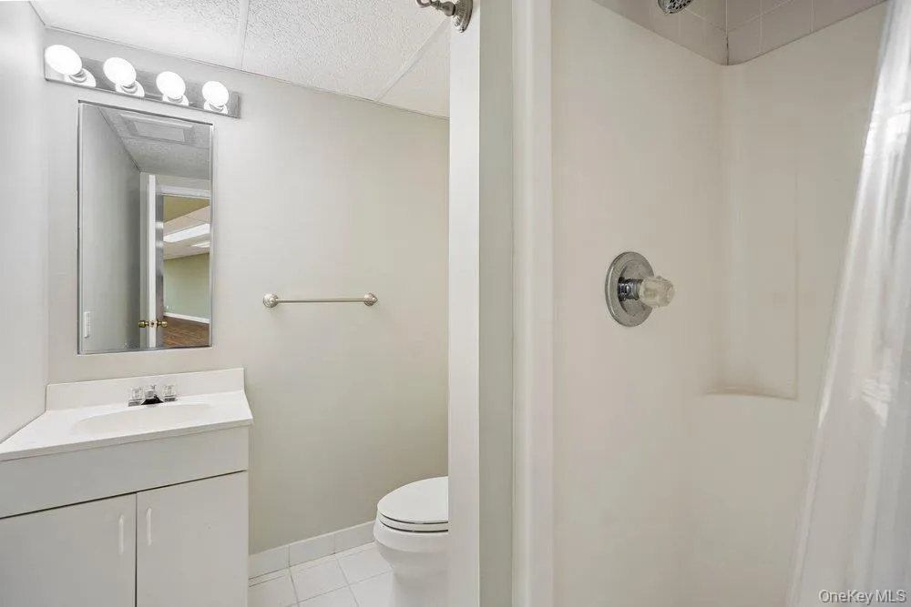 Bathroom featuring a white vanity with integrated sink, chrome faucet, white tile flooring, and a shower with a clear plastic curtain Bathroom featuring a white vanity with integrated sink, chrome faucet, white tile flooring, and a shower with a clear plastic curtain