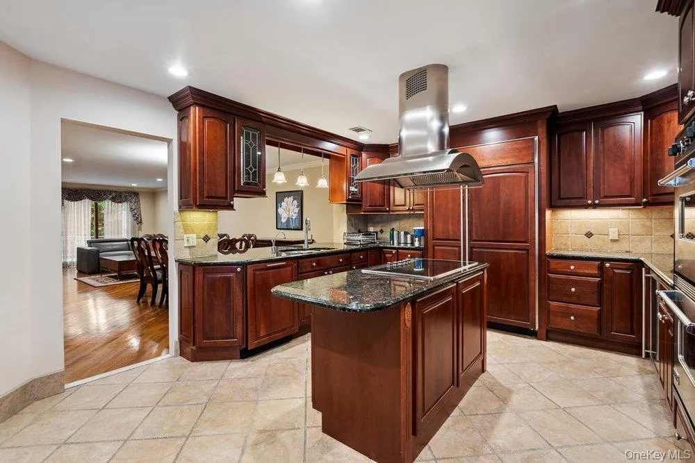Gourmet kitchen featuring rich wood cabinetry, granite countertops, a tiled backsplash, stainless steel appliances including a built-in oven and range hood, and a central island with a cooktop Gourmet kitchen featuring rich wood cabinetry, granite countertops, a tiled backsplash, stainless steel appliances including a built-in oven and range hood, and a central island with a cooktop
