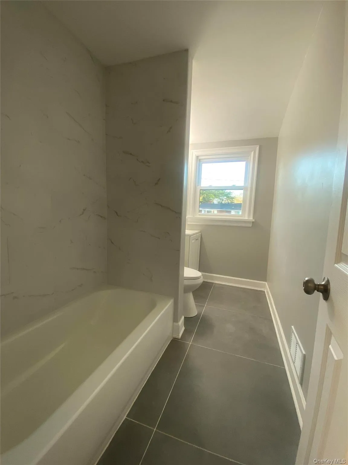 Bathroom featuring a white bathtub, white marble-look shower surround, dark gray tile flooring, and a window with white trim Bathroom featuring a white bathtub, white marble-look shower surround, dark gray tile flooring, and a window with white trim