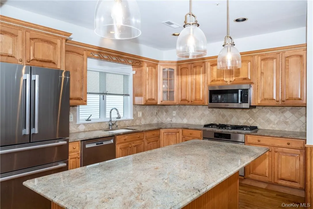 Kitchen featuring light wood cabinetry with crown molding, stainless steel appliances, a tiled backsplash, and a central island with a light-colored stone countertop Kitchen featuring light wood cabinetry with crown molding, stainless steel appliances, a tiled backsplash, and a central island with a light-colored stone countertop