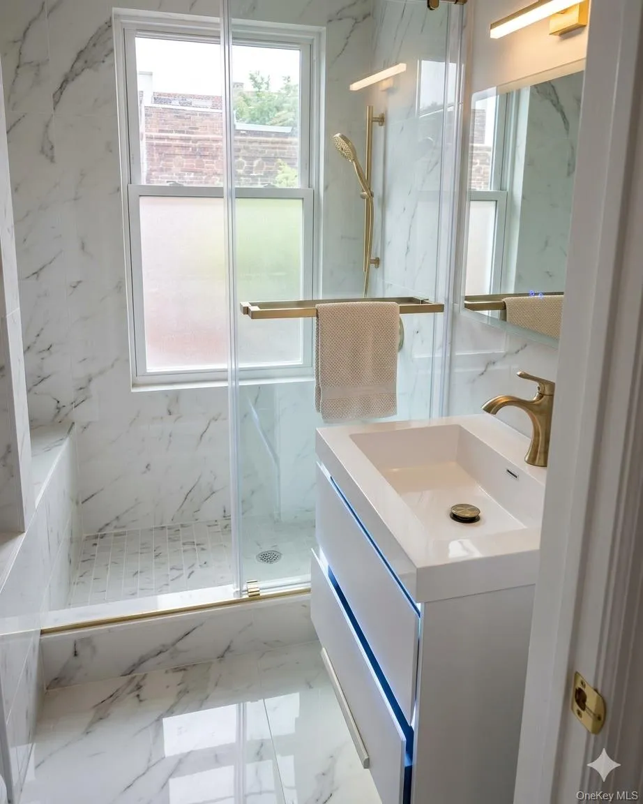 Bathroom featuring white marble-look tile, a frameless glass shower enclosure with brass-finish fixtures, a floating vanity with integrated lighting, and a window with frosted lower panes Bathroom featuring white marble-look tile, a frameless glass shower enclosure with brass-finish fixtures, a floating vanity with integrated lighting, and a window with frosted lower panes
