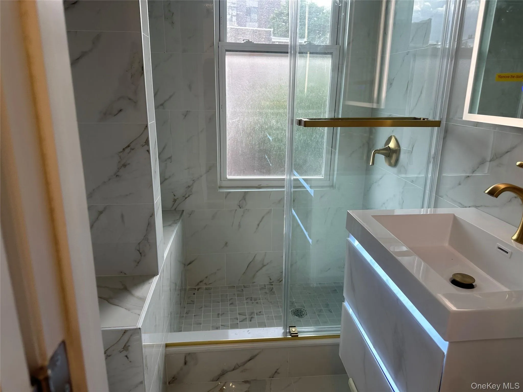 Bathroom featuring a glass-enclosed shower with a built-in bench, marble-look wall tiles, mosaic tile shower floor, and a modern vanity with integrated lighting and brass fixtures Bathroom featuring a glass-enclosed shower with a built-in bench, marble-look wall tiles, mosaic tile shower floor, and a modern vanity with integrated lighting and brass fixtures