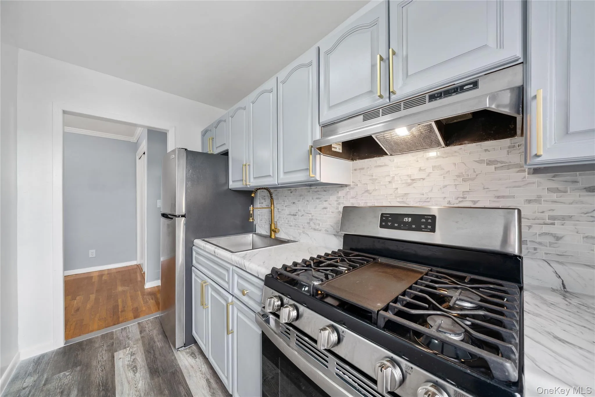 Modern kitchen featuring light gray cabinetry with gold-tone hardware, a stainless steel gas range with griddle, a stainless steel refrigerator, and a white marble-look countertop Modern kitchen featuring light gray cabinetry with gold-tone hardware, a stainless steel gas range with griddle, a stainless steel refrigerator, and a white marble-look countertop