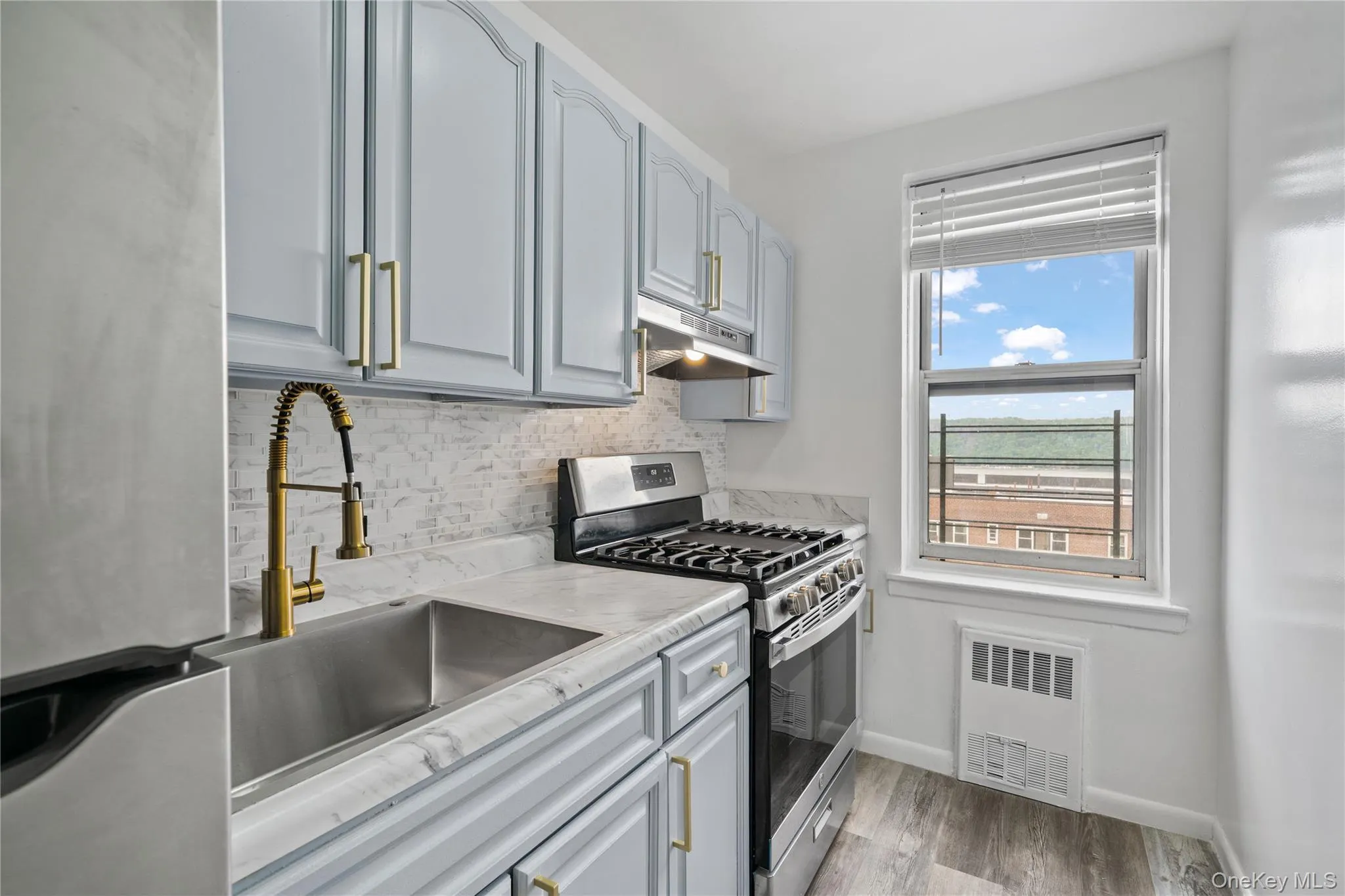 Kitchen featuring light blue cabinetry with gold-tone hardware, a stainless steel sink with a gold-tone pull-down faucet, a tile backsplash, and wood-finish flooring Kitchen featuring light blue cabinetry with gold-tone hardware, a stainless steel sink with a gold-tone pull-down faucet, a tile backsplash, and wood-finish flooring