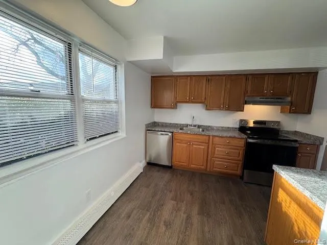 Kitchen with stainless steel appliances, dark wood-style flooring, a baseboard radiator, under cabinet range hood, and brown cabinetry Kitchen with stainless steel appliances, dark wood-style flooring, a baseboard radiator, under cabinet range hood, and brown cabinetry