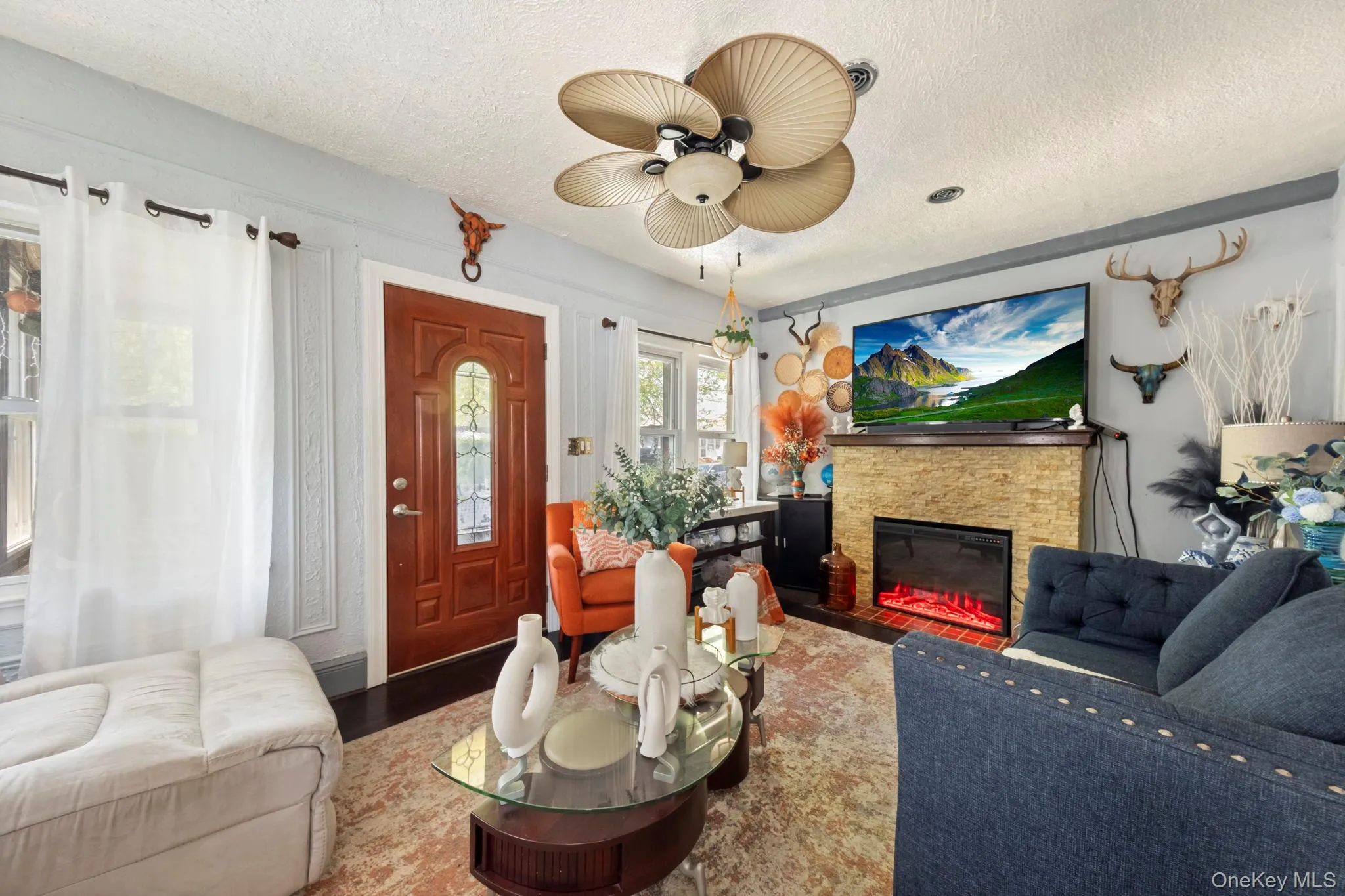 Living room featuring a textured ceiling, a ceiling fan with leaf-shaped blades, and a stained wood entry door with an arched glass insert Living room featuring a textured ceiling, a ceiling fan with leaf-shaped blades, and a stained wood entry door with an arched glass insert