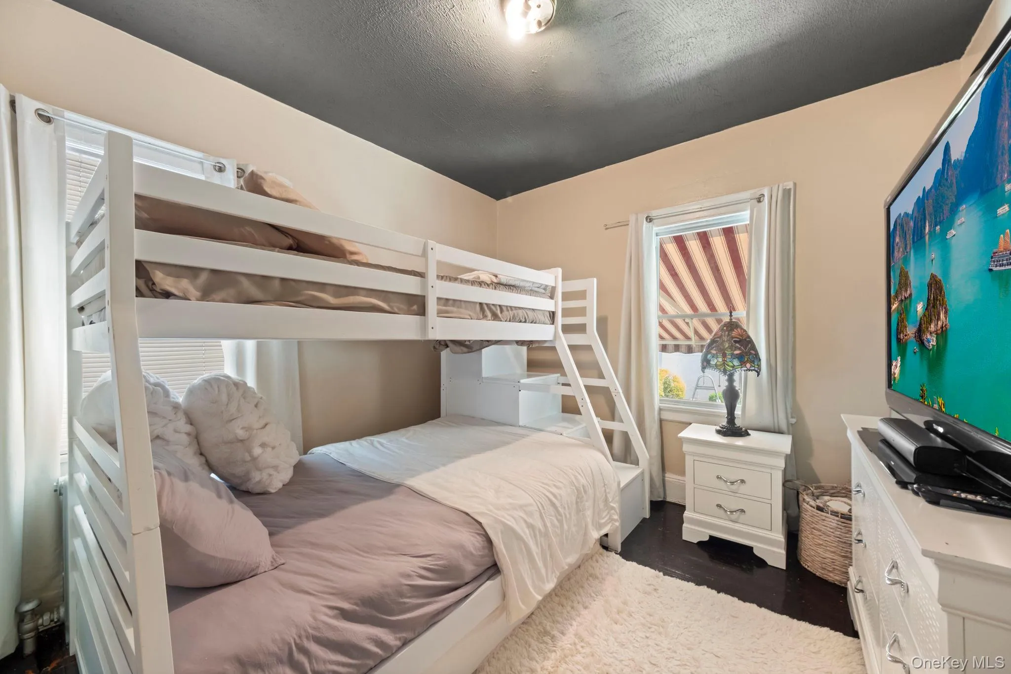 Bedroom featuring a white bunk bed with integrated steps, dark wood-finish flooring, and a window with blinds and drapes Bedroom featuring a white bunk bed with integrated steps, dark wood-finish flooring, and a window with blinds and drapes
