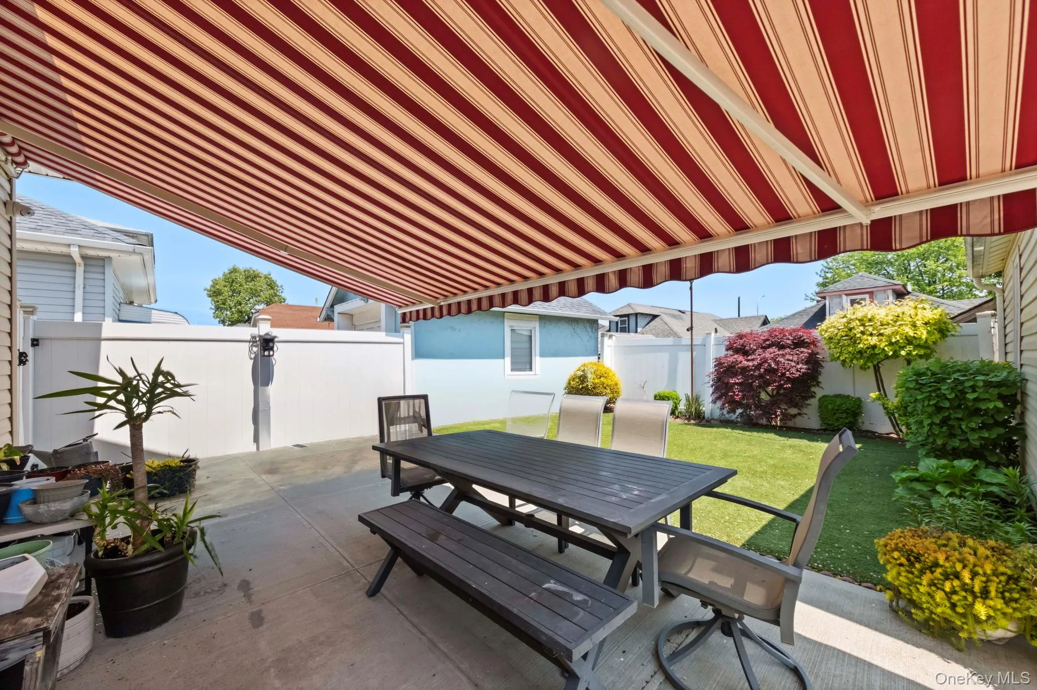 Patio area featuring a red and beige striped retractable awning, concrete flooring, white privacy fencing, and a manicured lawn with mature landscaping Patio area featuring a red and beige striped retractable awning, concrete flooring, white privacy fencing, and a manicured lawn with mature landscaping