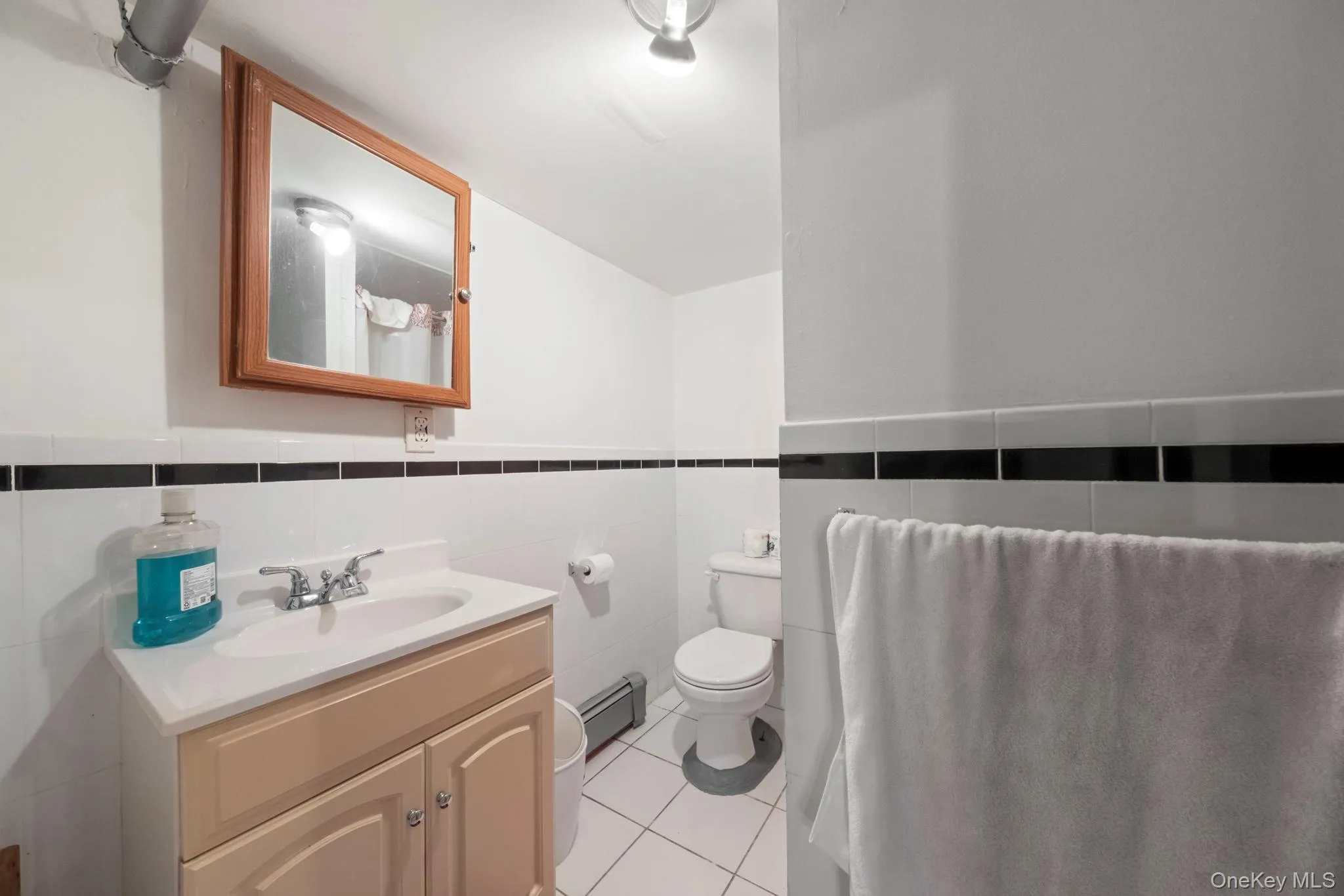 Downstairs bathroom featuring a single vanity with base cabinetry, tiled flooring, a wall-mounted medicine cabinet, and a tiled lower wall with a contrasting black accent band Downstairs bathroom featuring a single vanity with base cabinetry, tiled flooring, a wall-mounted medicine cabinet, and a tiled lower wall with a contrasting black accent band