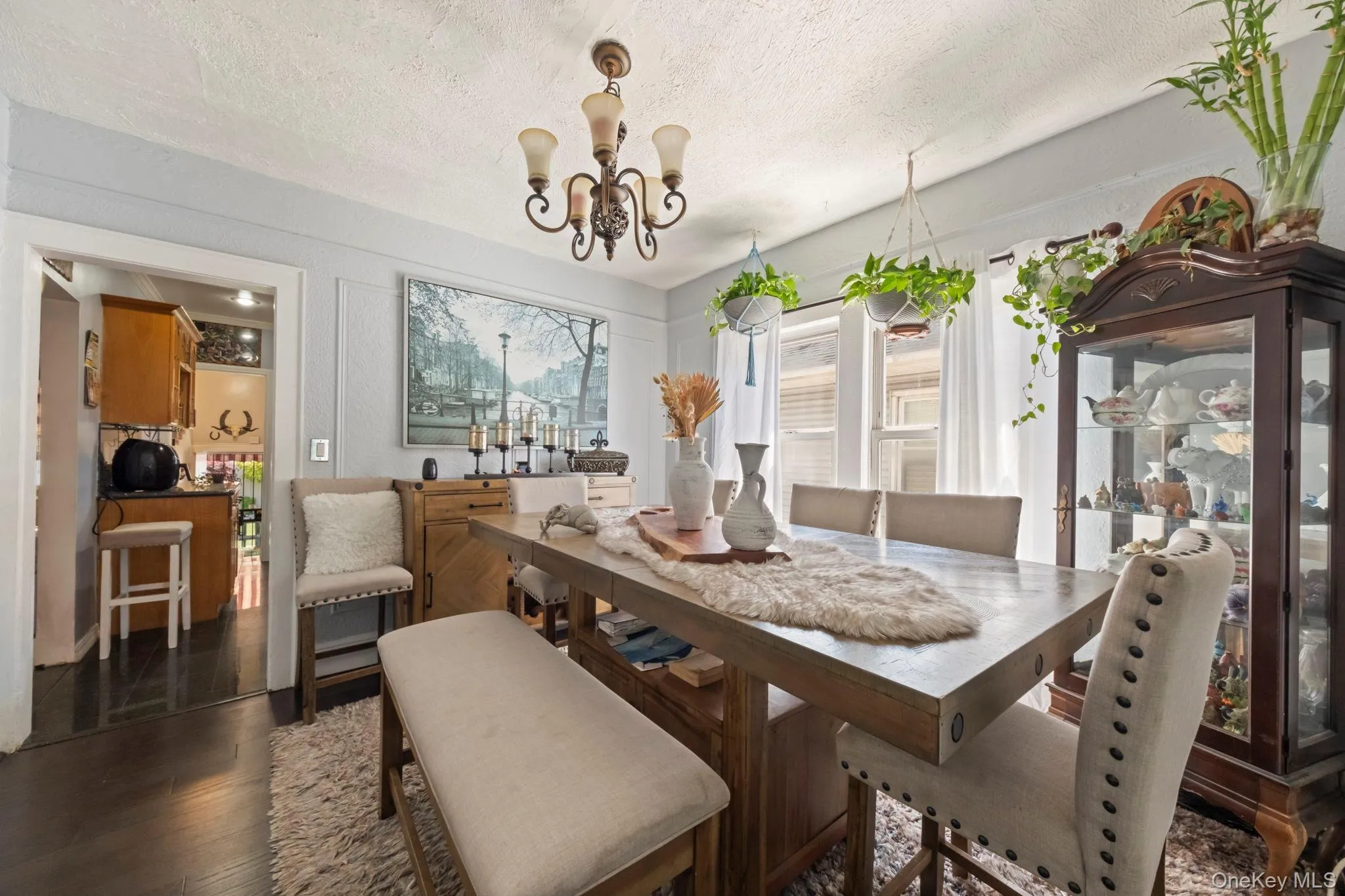 Dining area featuring a wood-finish table, upholstered bench and chairs, a decorative chandelier, and three large windows with white curtains Dining area featuring a wood-finish table, upholstered bench and chairs, a decorative chandelier, and three large windows with white curtains