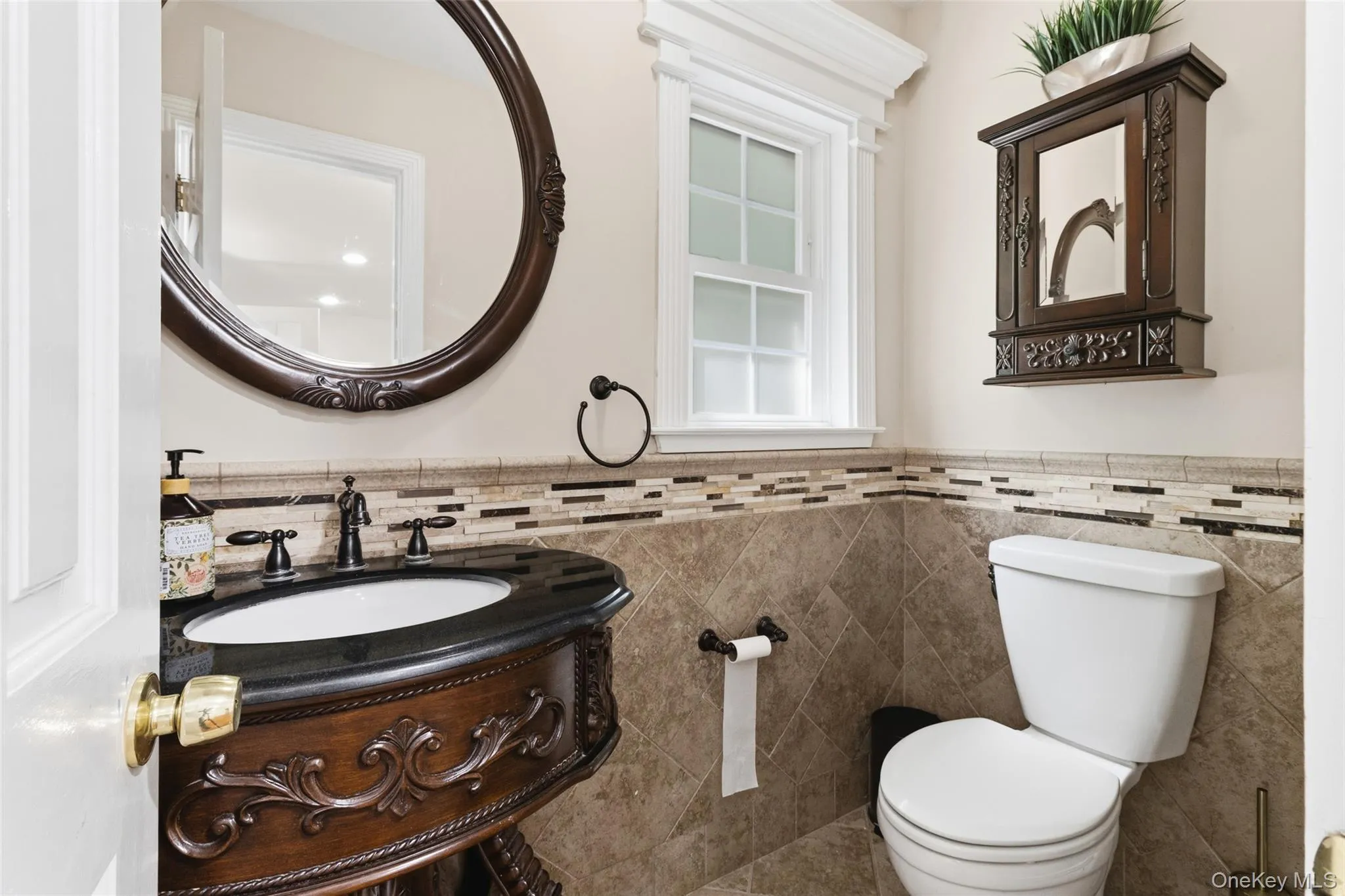 Powder room featuring a carved wood vanity with a black countertop, an oval framed mirror, an operable window with white trim, a decorative tile wainscoting, and large format floor tiling Powder room featuring a carved wood vanity with a black countertop, an oval framed mirror, an operable window with white trim, a decorative tile wainscoting, and large format floor tiling