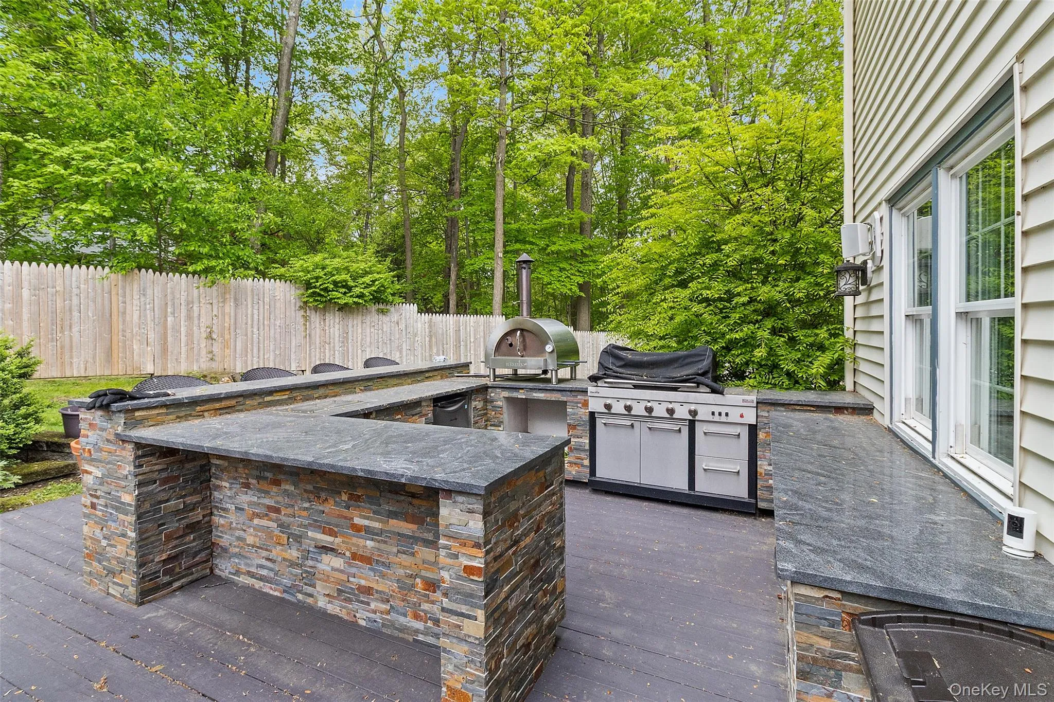 Outdoor kitchen featuring a U-shaped bar with stacked stone facade and dark countertops, a built-in grill, and an additional outdoor cooking appliance Outdoor kitchen featuring a U-shaped bar with stacked stone facade and dark countertops, a built-in grill, and an additional outdoor cooking appliance