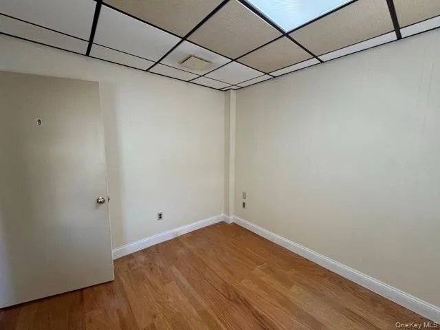 Room featuring wood-finish flooring, light-toned walls, and a dropped ceiling with integrated lighting panels Room featuring wood-finish flooring, light-toned walls, and a dropped ceiling with integrated lighting panels