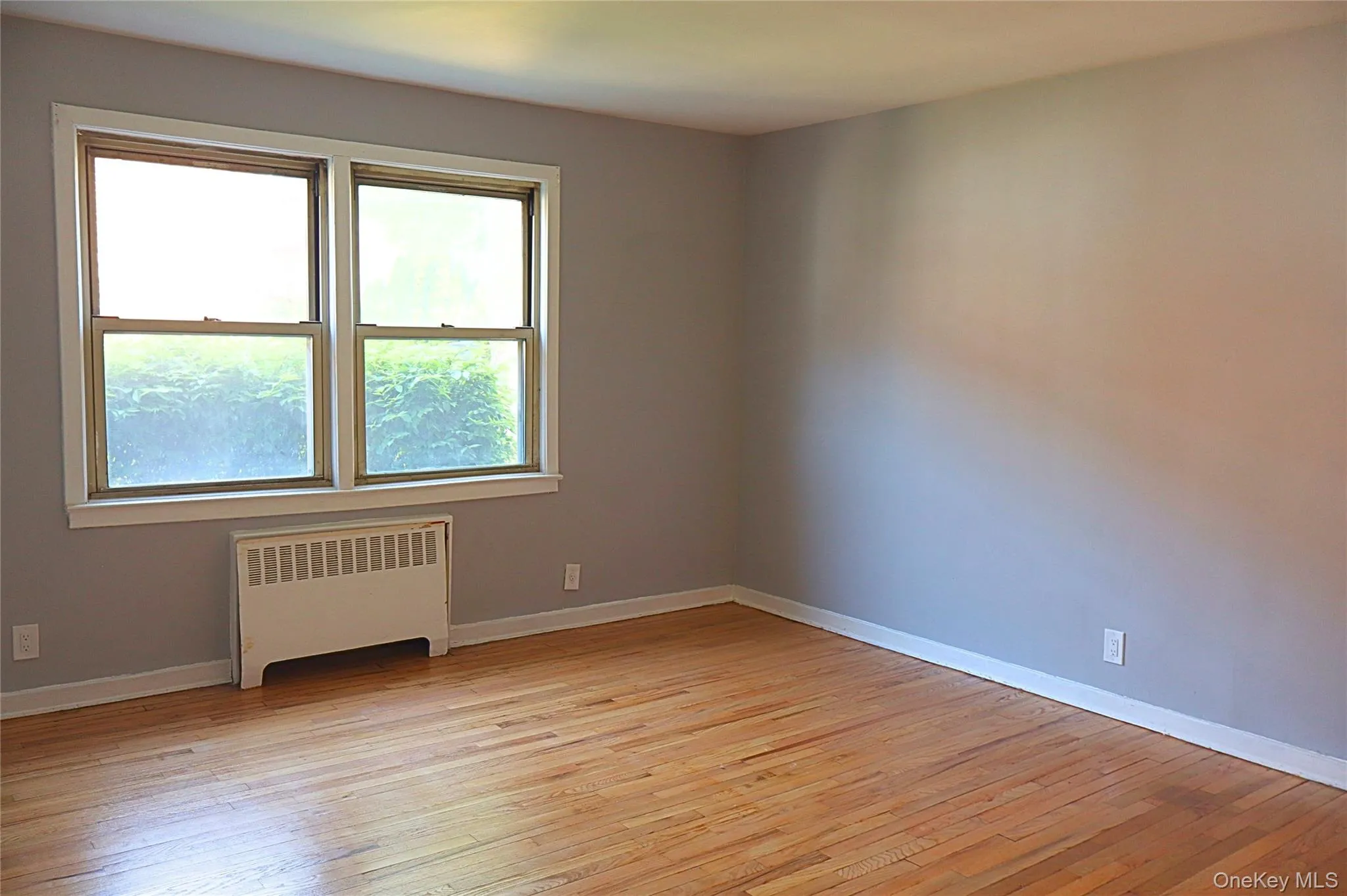Bedroom featuring light gray walls, wood-finish flooring, white baseboards, and a window unit with white trim Bedroom featuring light gray walls, wood-finish flooring, white baseboards, and a window unit with white trim