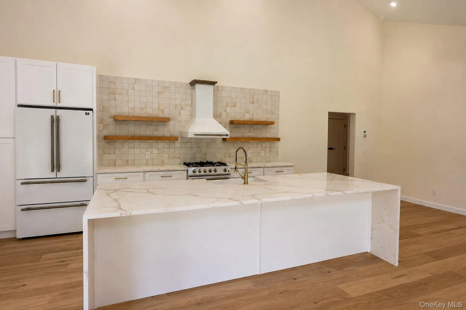 Kitchen featuring a waterfall-edge island with integrated sink, gas range, wood-finish flooring, white cabinetry, and a full-height tile backsplash with floating wood shelving Kitchen featuring a waterfall-edge island with integrated sink, gas range, wood-finish flooring, white cabinetry, and a full-height tile backsplash with floating wood shelving