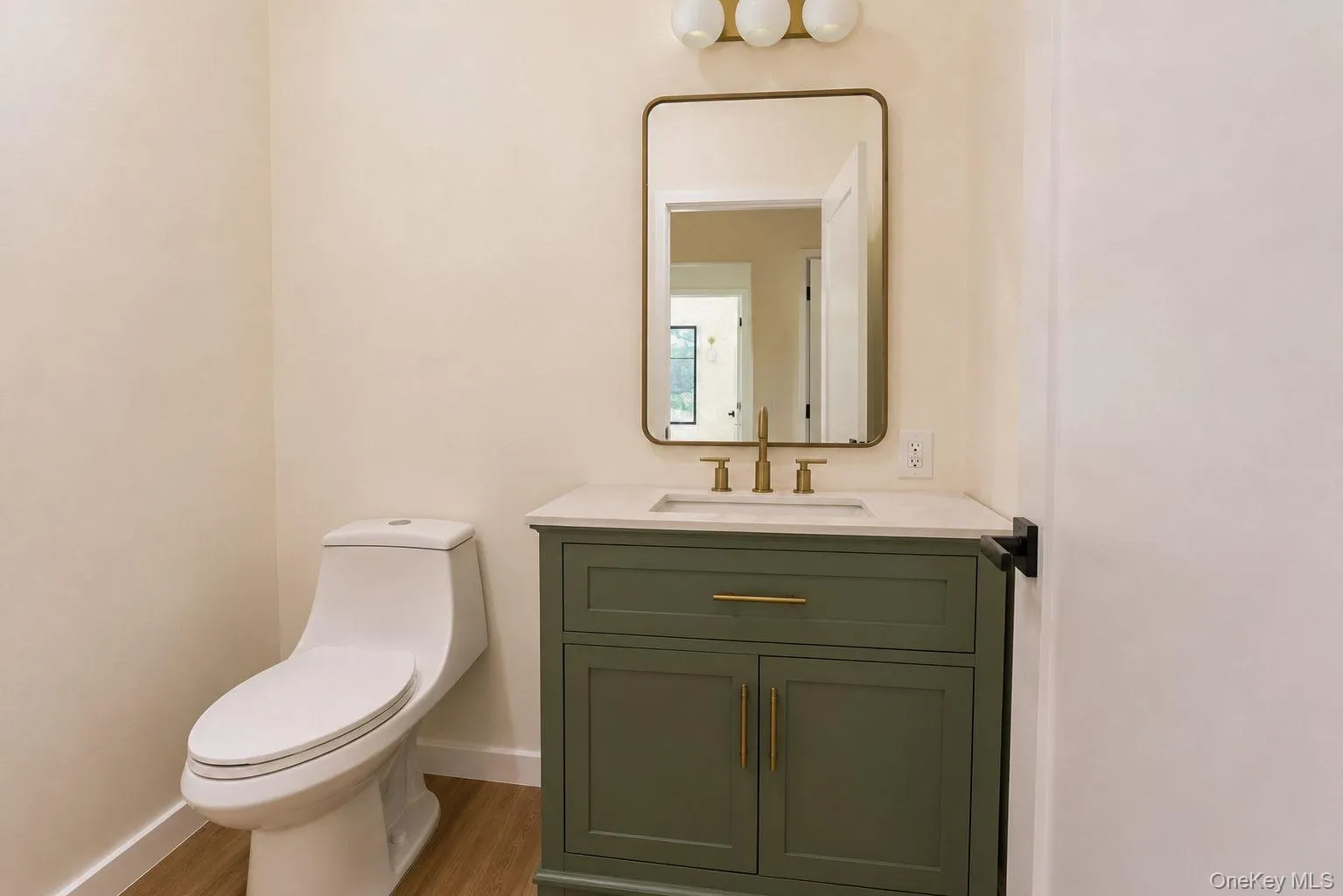 Powder room featuring a sage green vanity with a light-toned countertop, gold-finish hardware and faucet, a rectangular framed mirror, and wood-finish flooring Powder room featuring a sage green vanity with a light-toned countertop, gold-finish hardware and faucet, a rectangular framed mirror, and wood-finish flooring