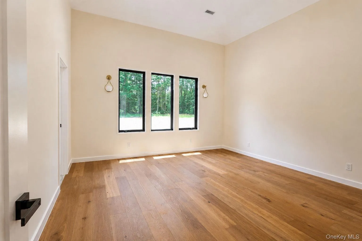 Spacious room featuring light-toned walls, wood-finish flooring, and three vertical windows with black frames Spacious room featuring light-toned walls, wood-finish flooring, and three vertical windows with black frames