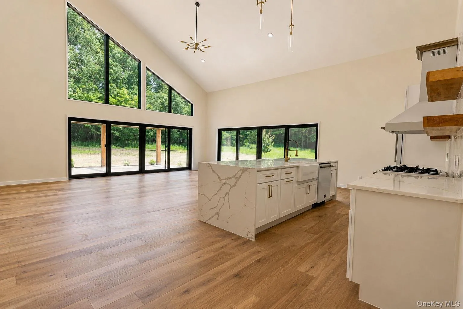 Vaulted ceiling great room featuring expansive windows, wood-finish flooring, a kitchen island with a waterfall countertop, and a built-in gas range with a stainless steel vent hood Vaulted ceiling great room featuring expansive windows, wood-finish flooring, a kitchen island with a waterfall countertop, and a built-in gas range with a stainless steel vent hood