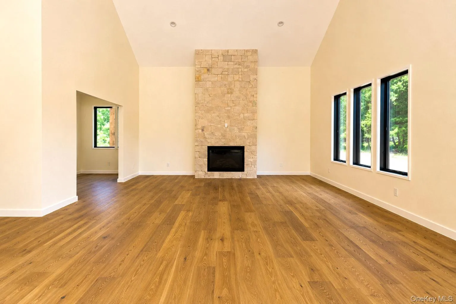 Spacious living area featuring a floor-to-ceiling stone fireplace, vaulted ceilings, natural wood-finish flooring, and multiple large windows with black frames. Access to office/den on left off the living room Spacious living area featuring a floor-to-ceiling stone fireplace, vaulted ceilings, natural wood-finish flooring, and multiple large windows with black frames. Access to office/den on left off the living room