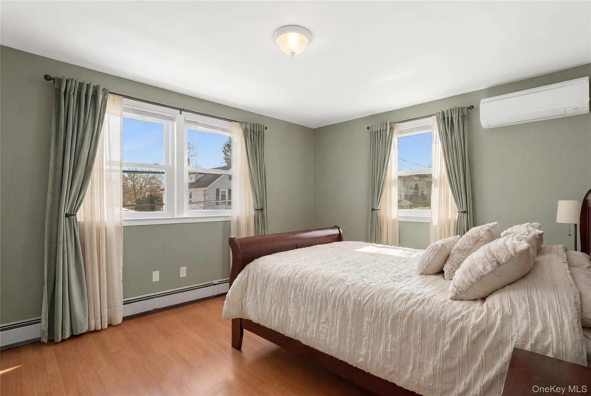 Bright bedroom featuring twin windows, wood-finish flooring, a wall-mounted climate control unit, and a ceiling-mounted light fixture Bright bedroom featuring twin windows, wood-finish flooring, a wall-mounted climate control unit, and a ceiling-mounted light fixture