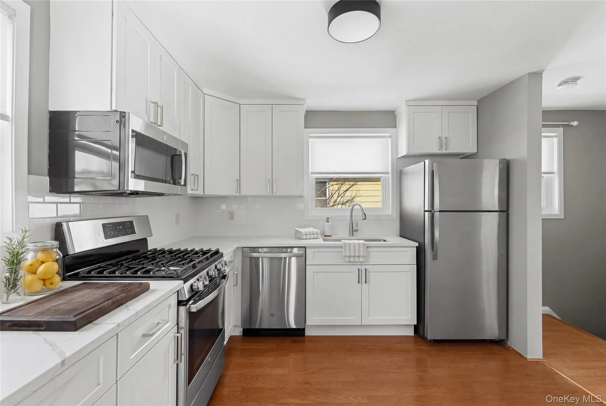 Modern kitchen featuring white shaker cabinetry, stainless steel appliances, white subway tile backsplash, light-toned countertops, and wood-finish flooring Modern kitchen featuring white shaker cabinetry, stainless steel appliances, white subway tile backsplash, light-toned countertops, and wood-finish flooring