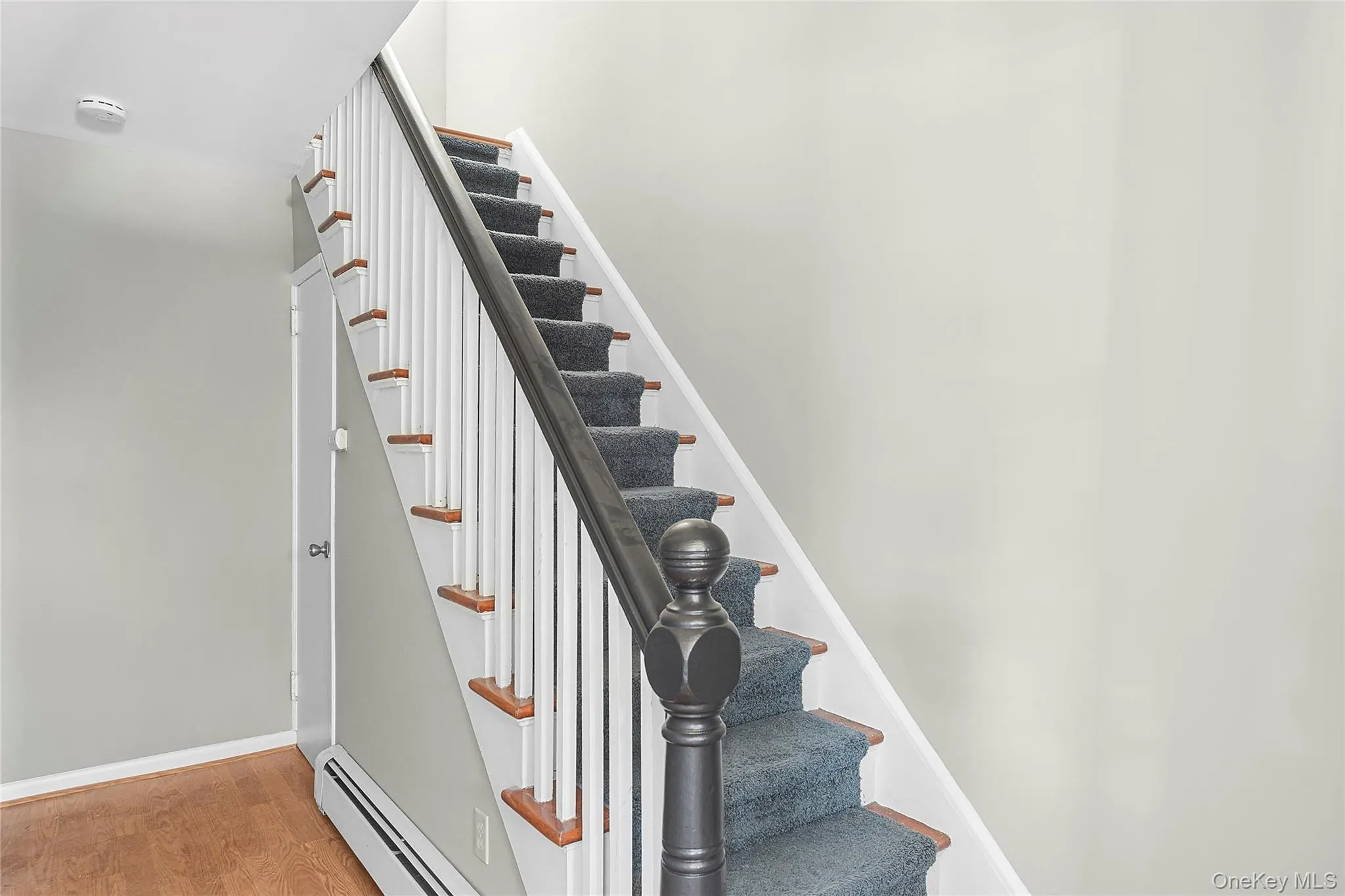 Staircase featuring dark carpeted treads, wood-finish risers, a white spindle railing, and a dark wood-finish newel post and handrail Staircase featuring dark carpeted treads, wood-finish risers, a white spindle railing, and a dark wood-finish newel post and handrail