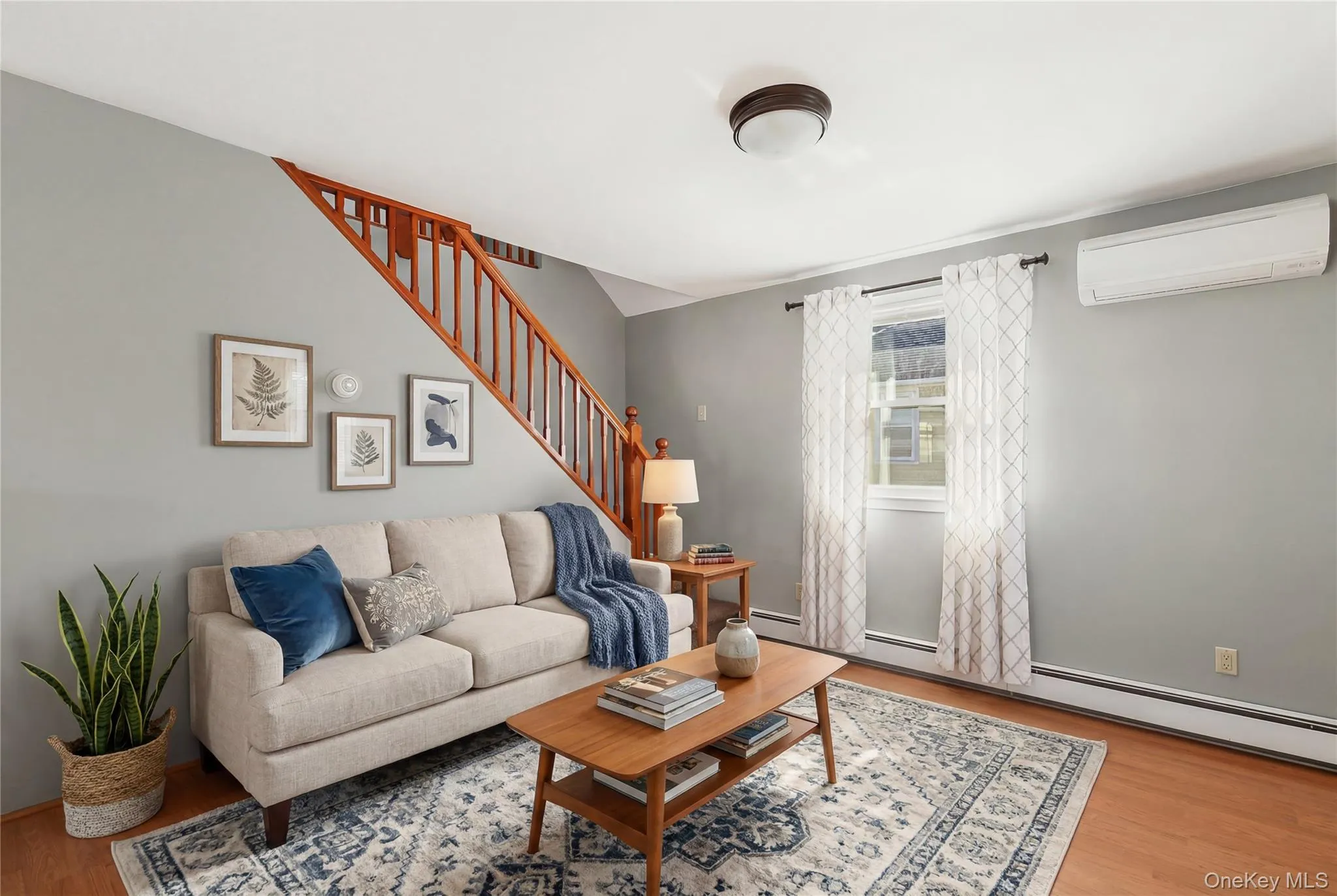 Living area with wood-finish flooring, a contemporary wall-mounted HVAC unit, and a prominent staircase with wood banister and spindles Living area with wood-finish flooring, a contemporary wall-mounted HVAC unit, and a prominent staircase with wood banister and spindles
