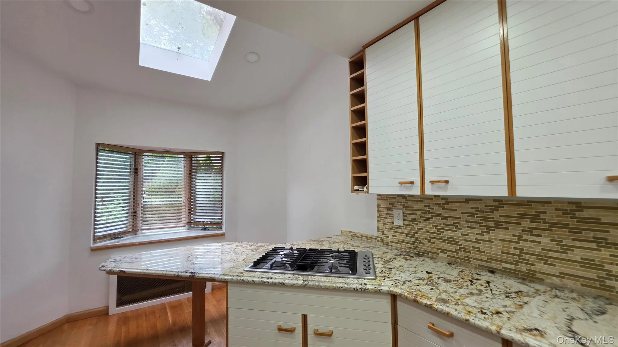 Kitchen featuring granite countertops, integrated gas cooktop, tile backsplash, white cabinetry with wood trim, and a skylight Kitchen featuring granite countertops, integrated gas cooktop, tile backsplash, white cabinetry with wood trim, and a skylight