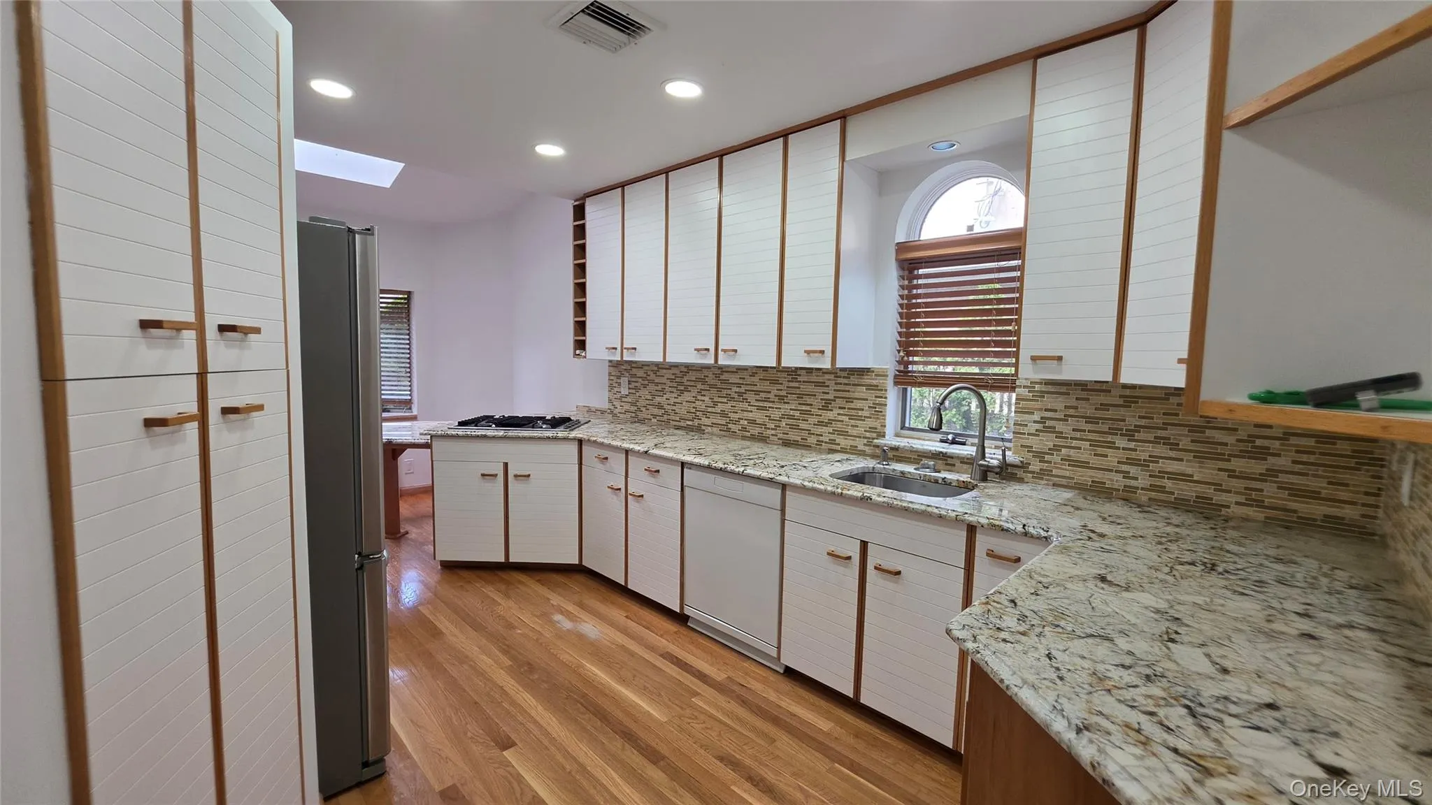 Modern kitchen featuring extensive white cabinetry with wood accents, light-toned granite countertops, a mosaic tile backsplash, and wood-finish flooring Modern kitchen featuring extensive white cabinetry with wood accents, light-toned granite countertops, a mosaic tile backsplash, and wood-finish flooring