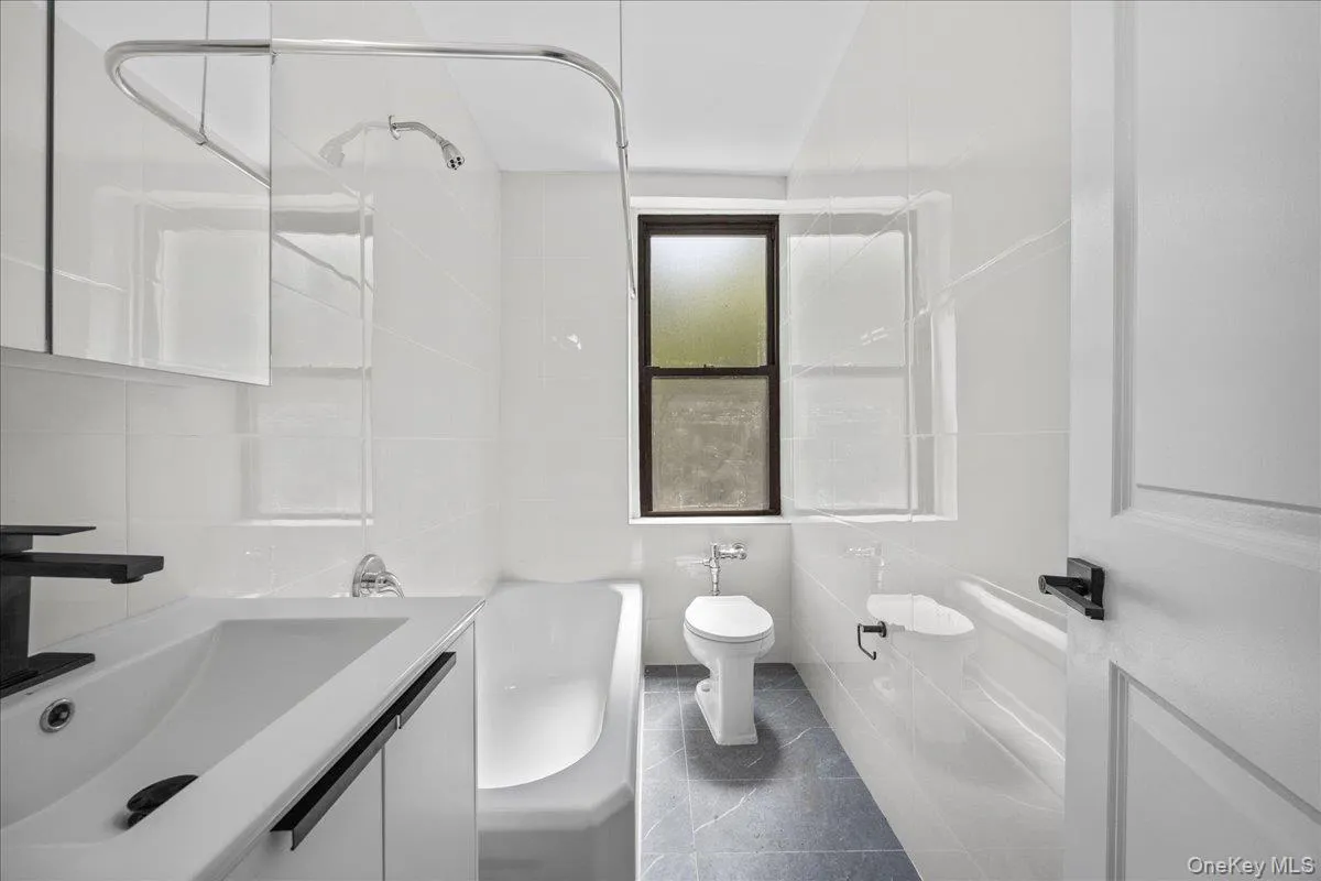 Modern bathroom featuring a white vanity with integrated sink, black fixtures, a bathtub with shower, a toilet, and a window with dark framing Modern bathroom featuring a white vanity with integrated sink, black fixtures, a bathtub with shower, a toilet, and a window with dark framing