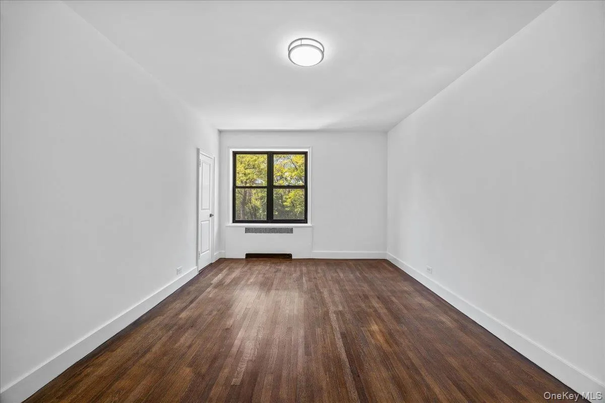 Spacious room featuring rich hardwood flooring, crisp white walls, and a large window with exterior foliage Spacious room featuring rich hardwood flooring, crisp white walls, and a large window with exterior foliage