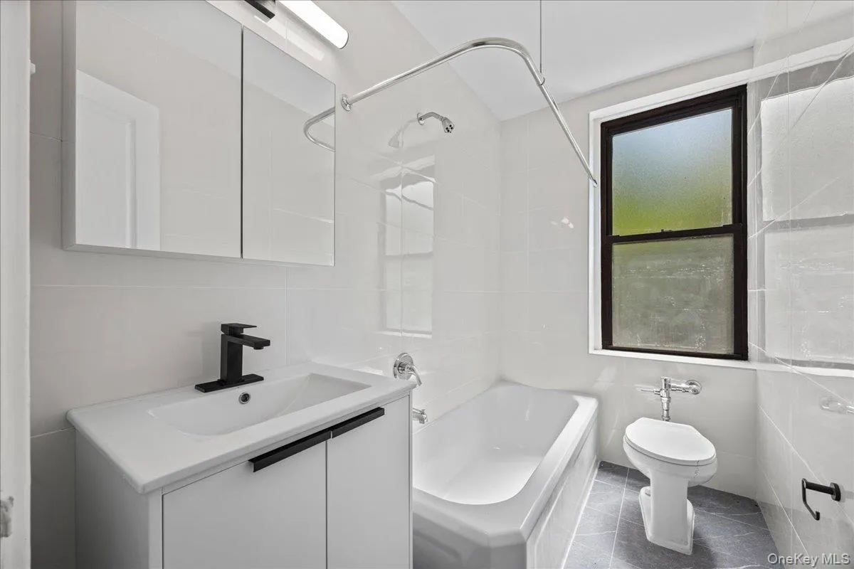 Modern bathroom featuring a white vanity with integrated sink, black fixtures, a bathtub with shower, a toilet, and a window with dark framing Modern bathroom featuring a white vanity with integrated sink, black fixtures, a bathtub with shower, a toilet, and a window with dark framing