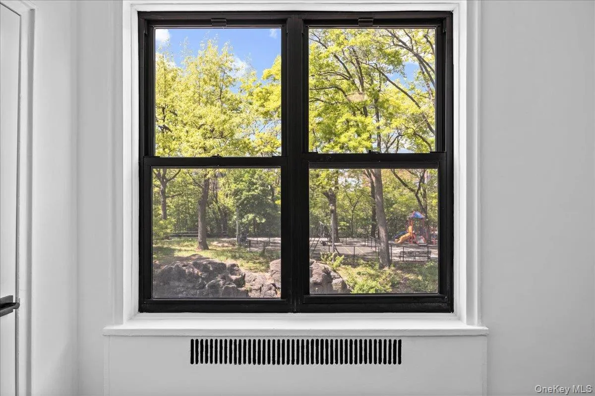 Black-framed double-hung window offering park scenery with mature trees and a distant play area, complemented by a white radiator cover and light-colored walls Black-framed double-hung window offering park scenery with mature trees and a distant play area, complemented by a white radiator cover and light-colored walls