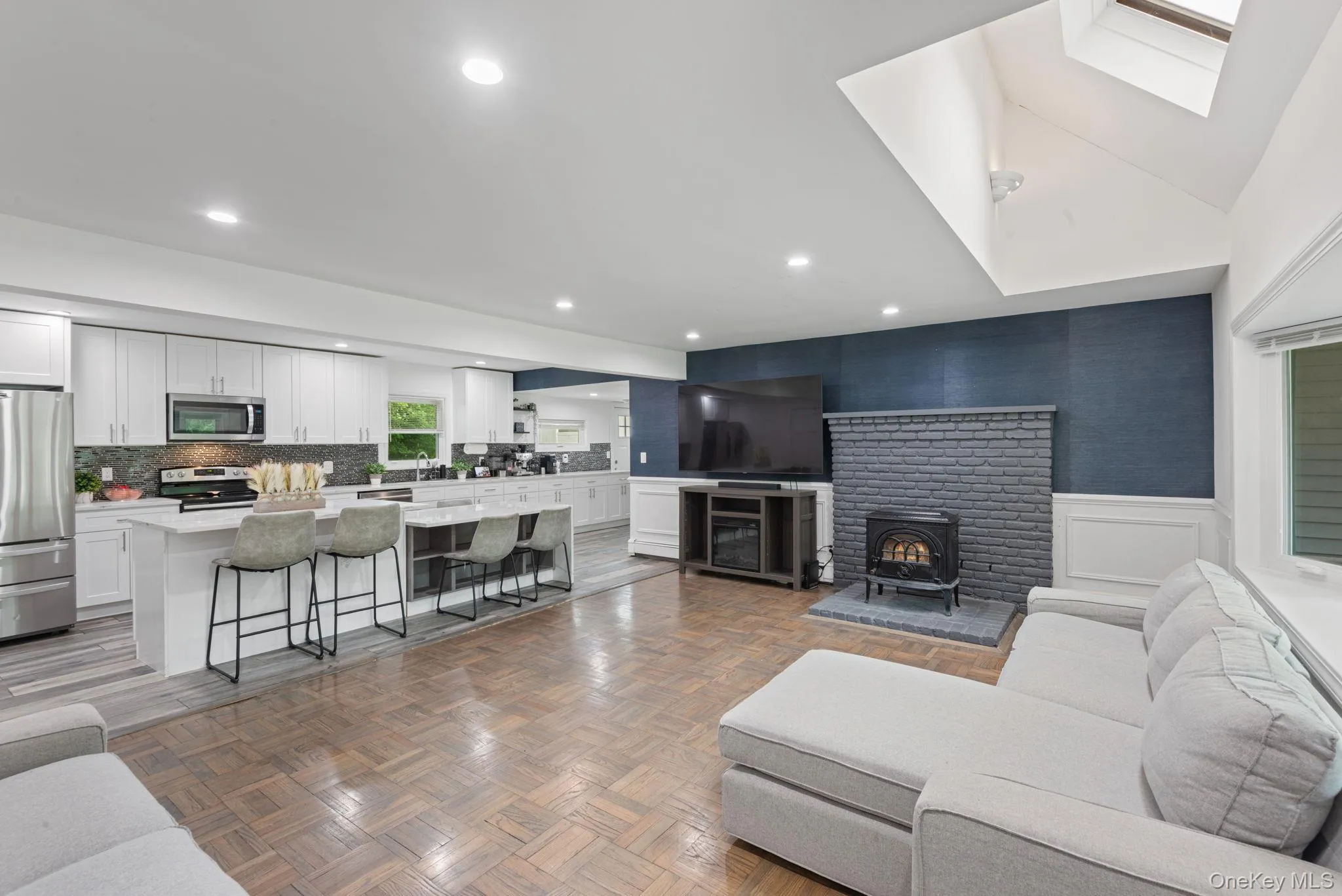 Living area featuring wood-finish parquet flooring, a bay window, recessed lighting, and white wainscoting with view into open-concept kitchen Living area featuring wood-finish parquet flooring, a bay window, recessed lighting, and white wainscoting with view into open-concept kitchen
