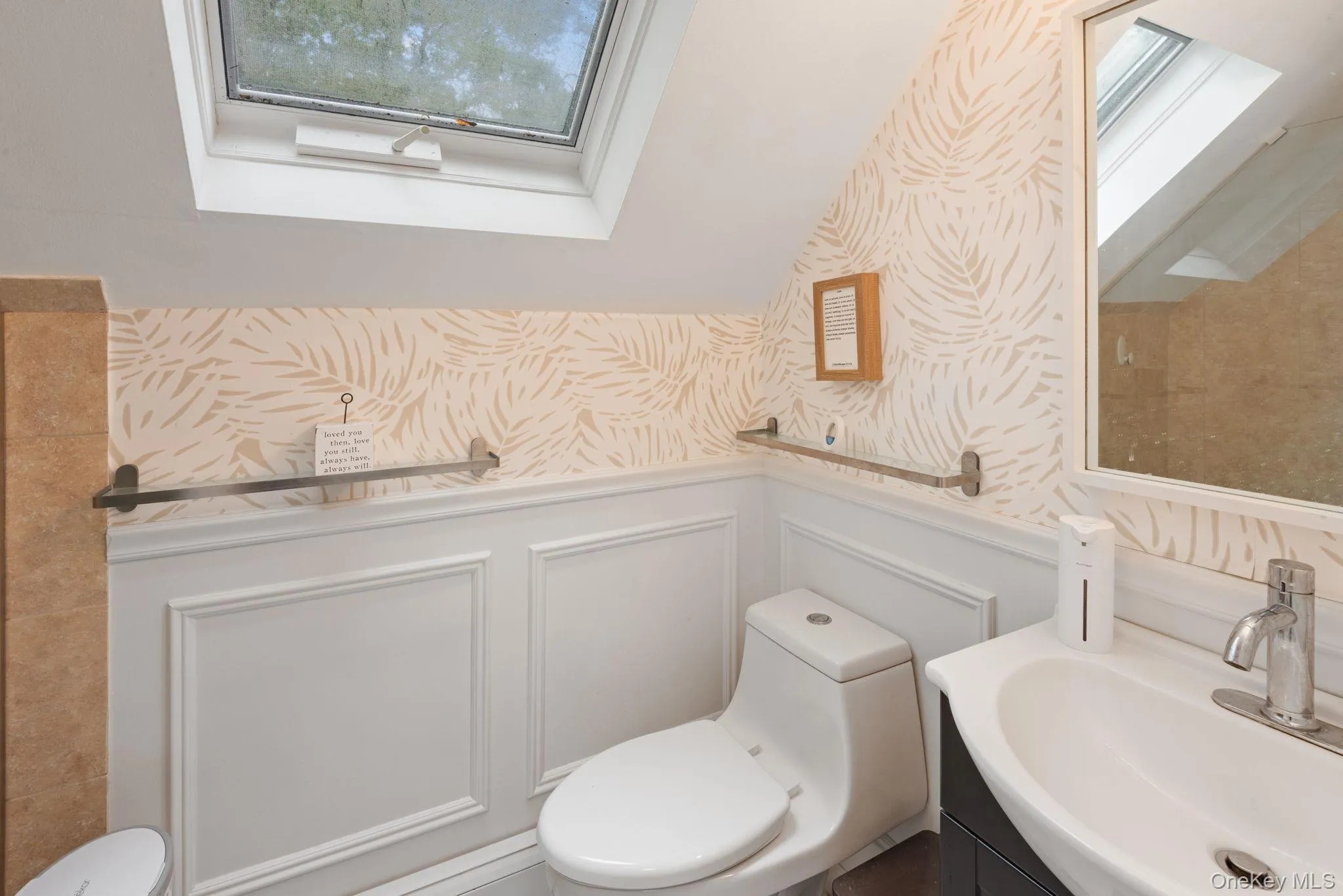 Ensuite bathroom featuring a skylight, modern toilet, vanity with a white basin and chrome faucet, wainscoting, and patterned wallpaper Ensuite bathroom featuring a skylight, modern toilet, vanity with a white basin and chrome faucet, wainscoting, and patterned wallpaper