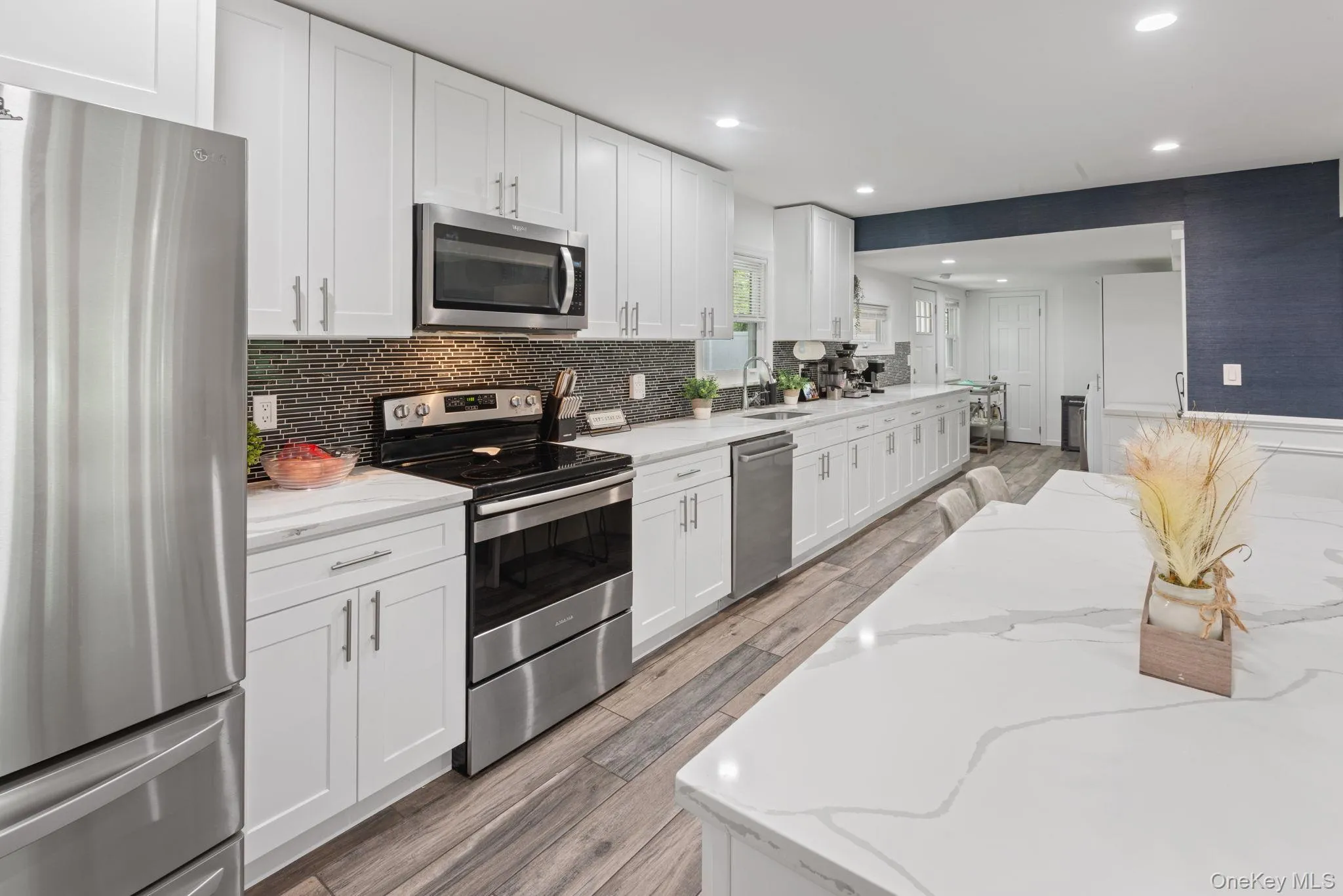 Modern kitchen featuring stainless steel appliances, white shaker cabinetry, white countertops, and a dark mosaic tile backsplash Modern kitchen featuring stainless steel appliances, white shaker cabinetry, white countertops, and a dark mosaic tile backsplash
