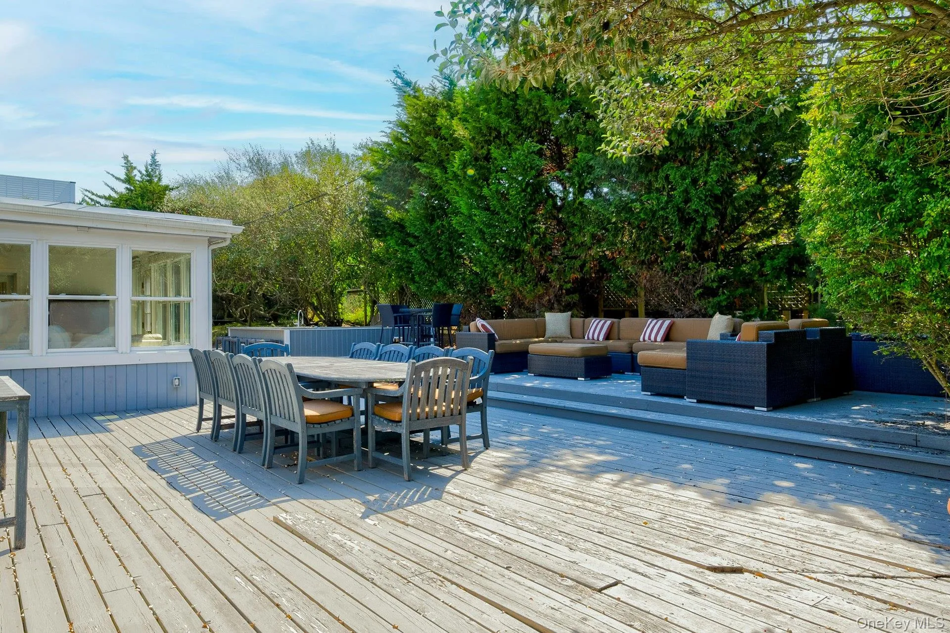 Multi-level wood deck featuring a dining area, a defined lounge space, and mature perimeter landscaping Multi-level wood deck featuring a dining area, a defined lounge space, and mature perimeter landscaping