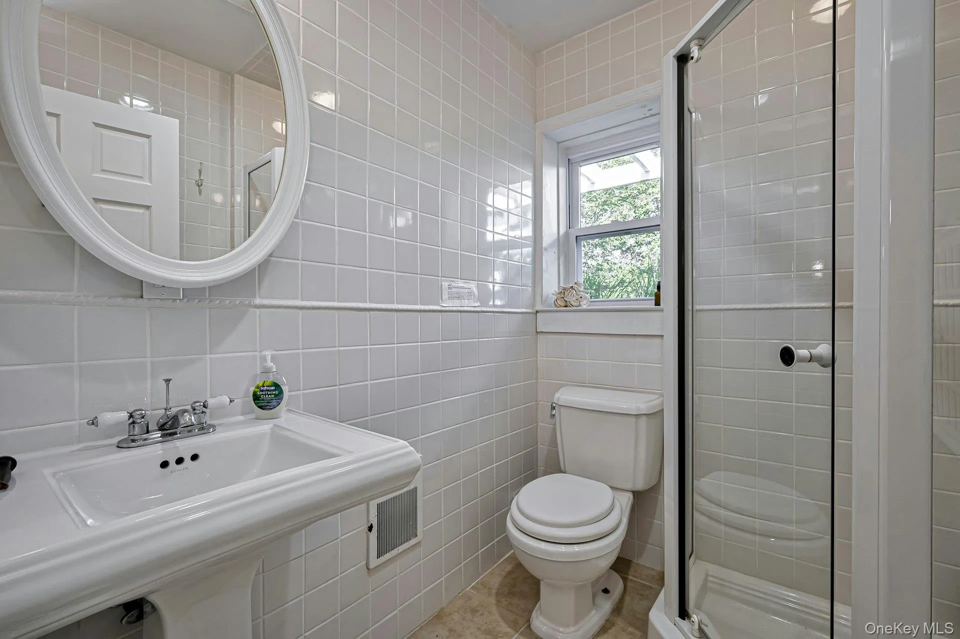 Bathroom featuring a pedestal sink, oval framed mirror, tiled walls, and a glass-enclosed shower Bathroom featuring a pedestal sink, oval framed mirror, tiled walls, and a glass-enclosed shower
