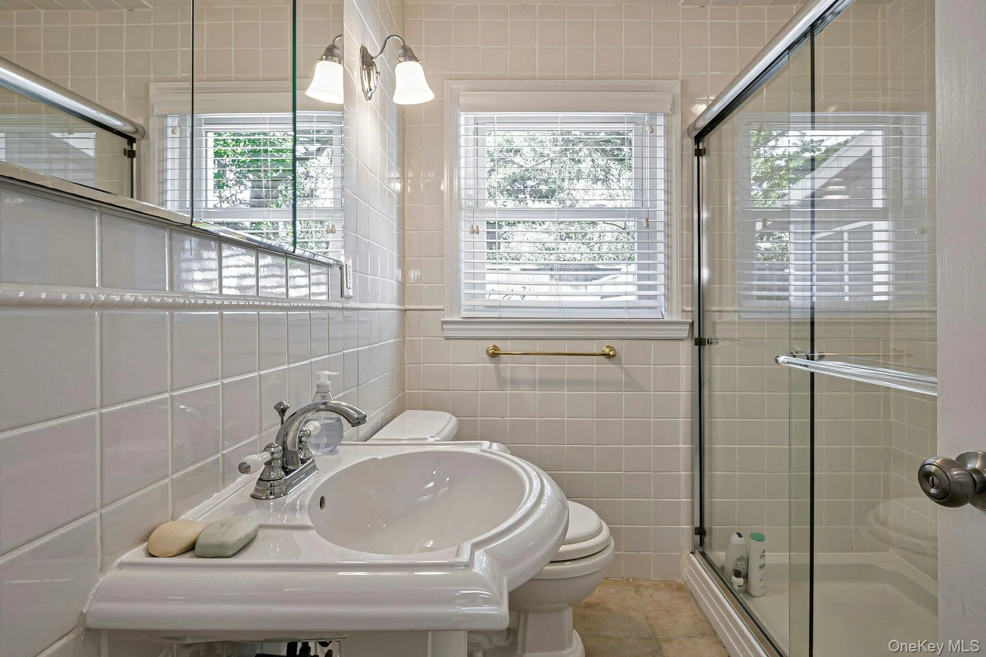 Bathroom featuring a pedestal sink, white tile walls, a sliding glass shower enclosure, and two windows with blinds Bathroom featuring a pedestal sink, white tile walls, a sliding glass shower enclosure, and two windows with blinds