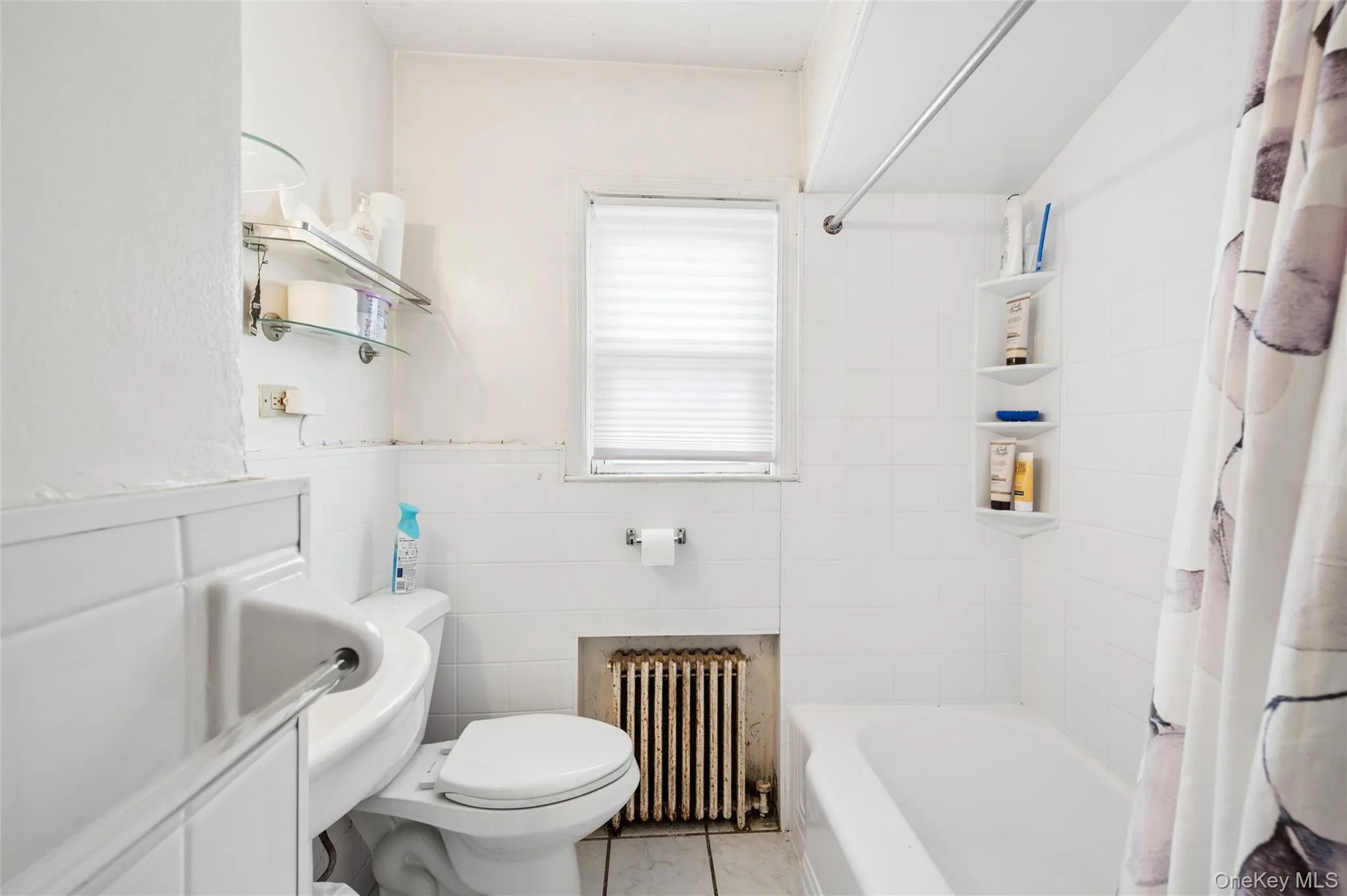 Bathroom featuring a white bathtub with a shower rod, a toilet, a window with blinds, and white wall tiling Bathroom featuring a white bathtub with a shower rod, a toilet, a window with blinds, and white wall tiling