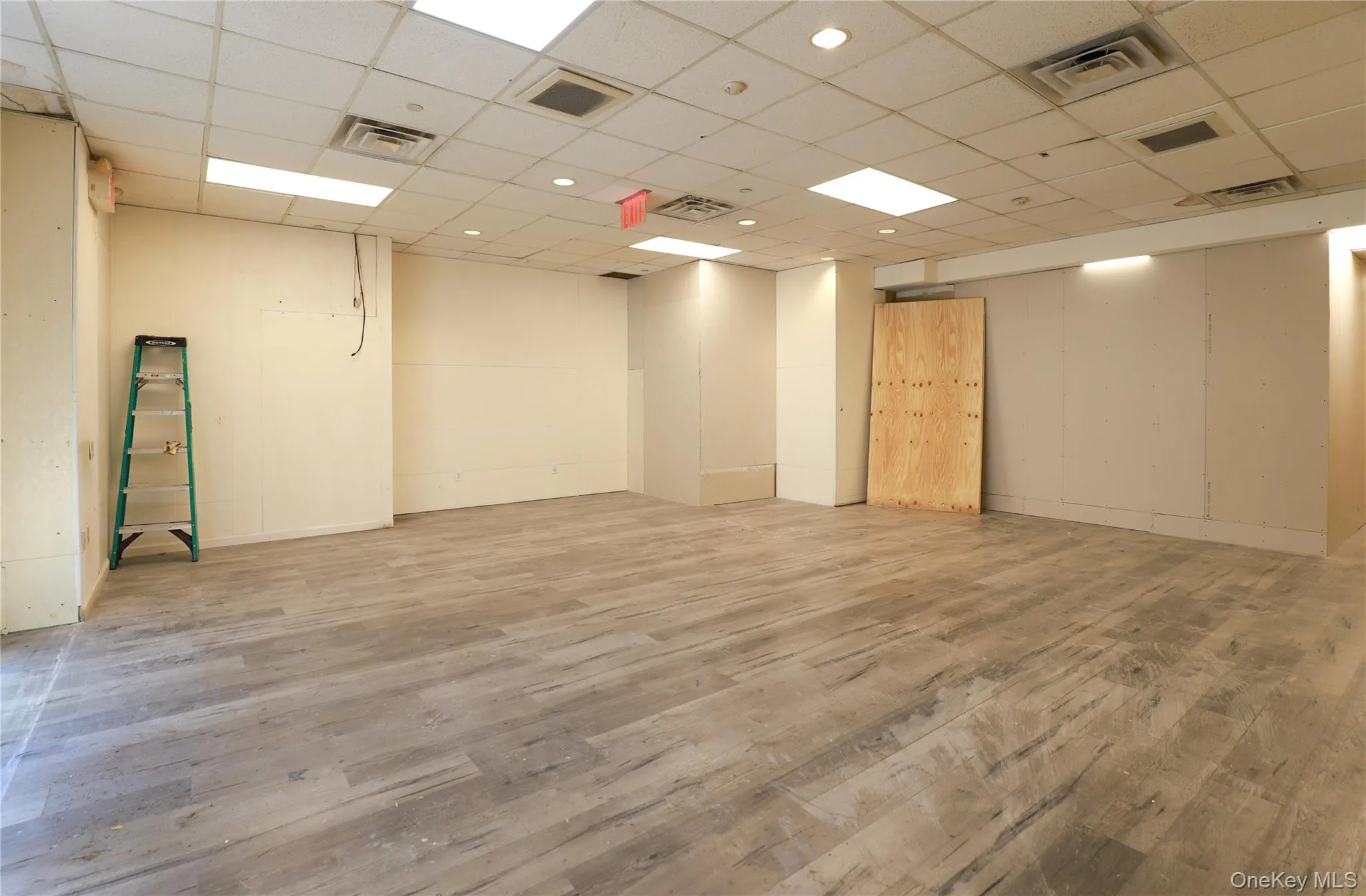 Spacious room featuring wood-finish flooring, drop ceiling with recessed lighting, and neutral wall tones Spacious room featuring wood-finish flooring, drop ceiling with recessed lighting, and neutral wall tones