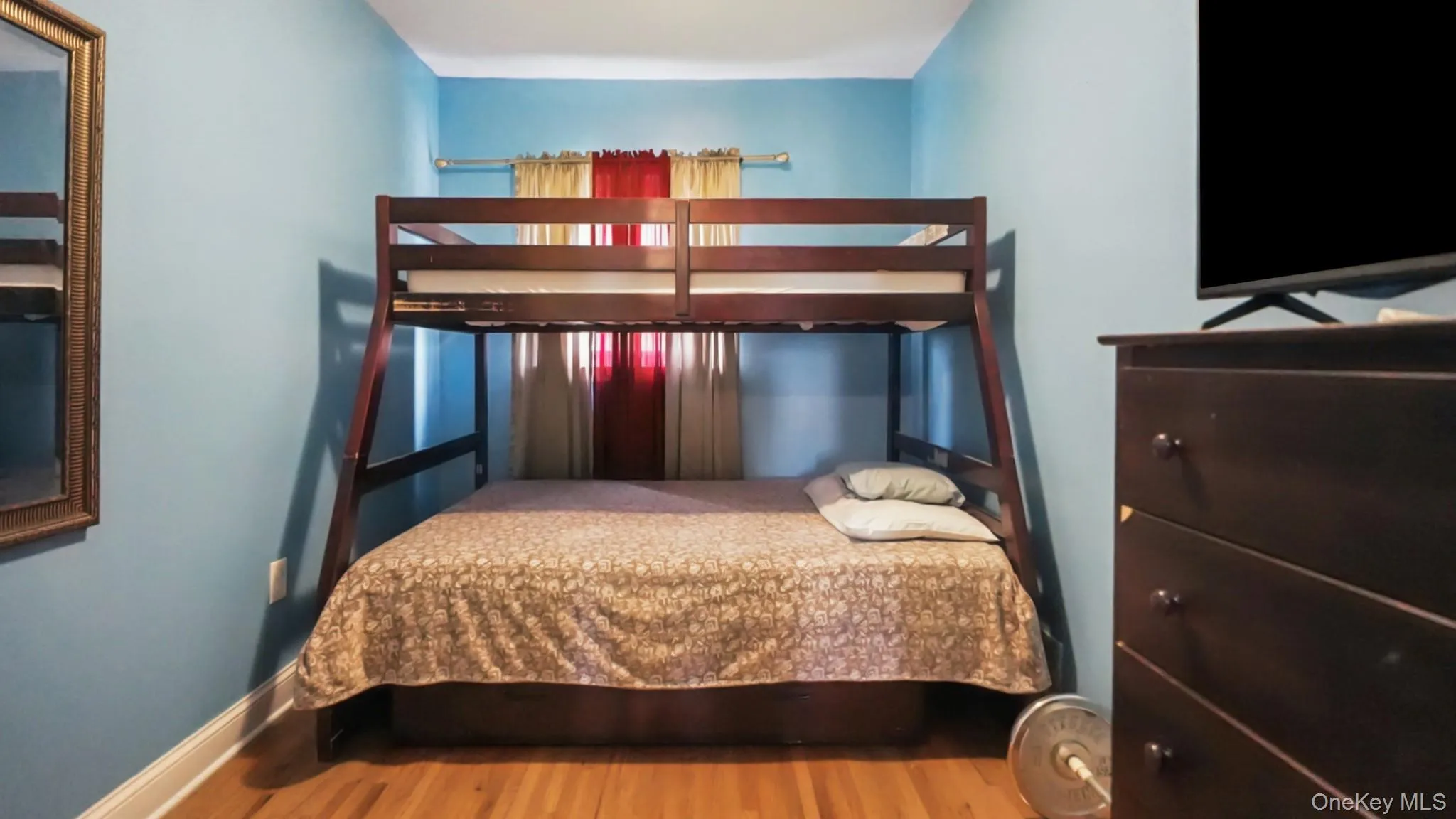 Bedroom featuring light blue walls, wood-finish flooring, and a window with red and white curtain panels Bedroom featuring light blue walls, wood-finish flooring, and a window with red and white curtain panels
