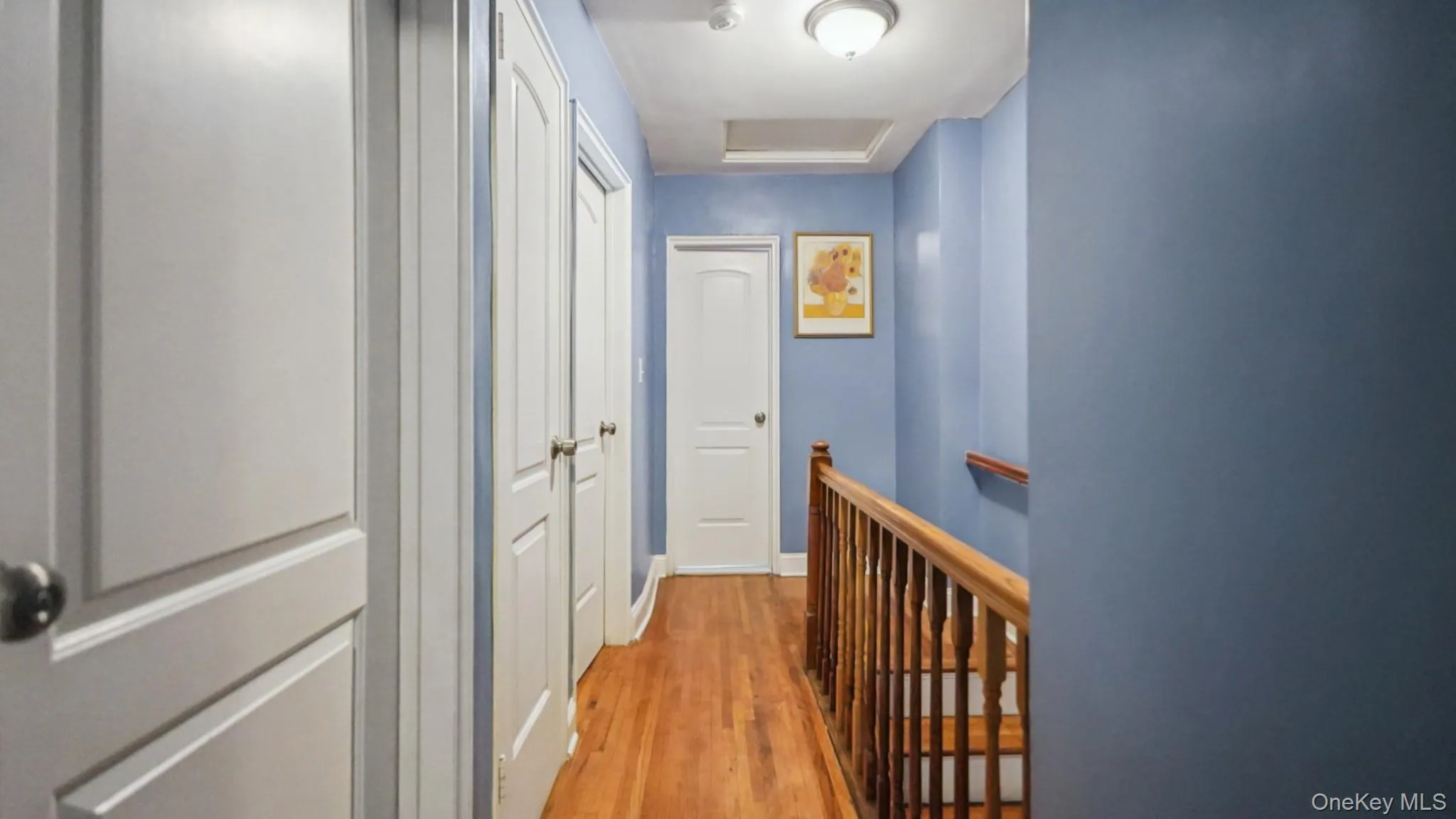 Hallway featuring wood-finish flooring, white panel doors, wood stair railing, and a flush mount ceiling light Hallway featuring wood-finish flooring, white panel doors, wood stair railing, and a flush mount ceiling light