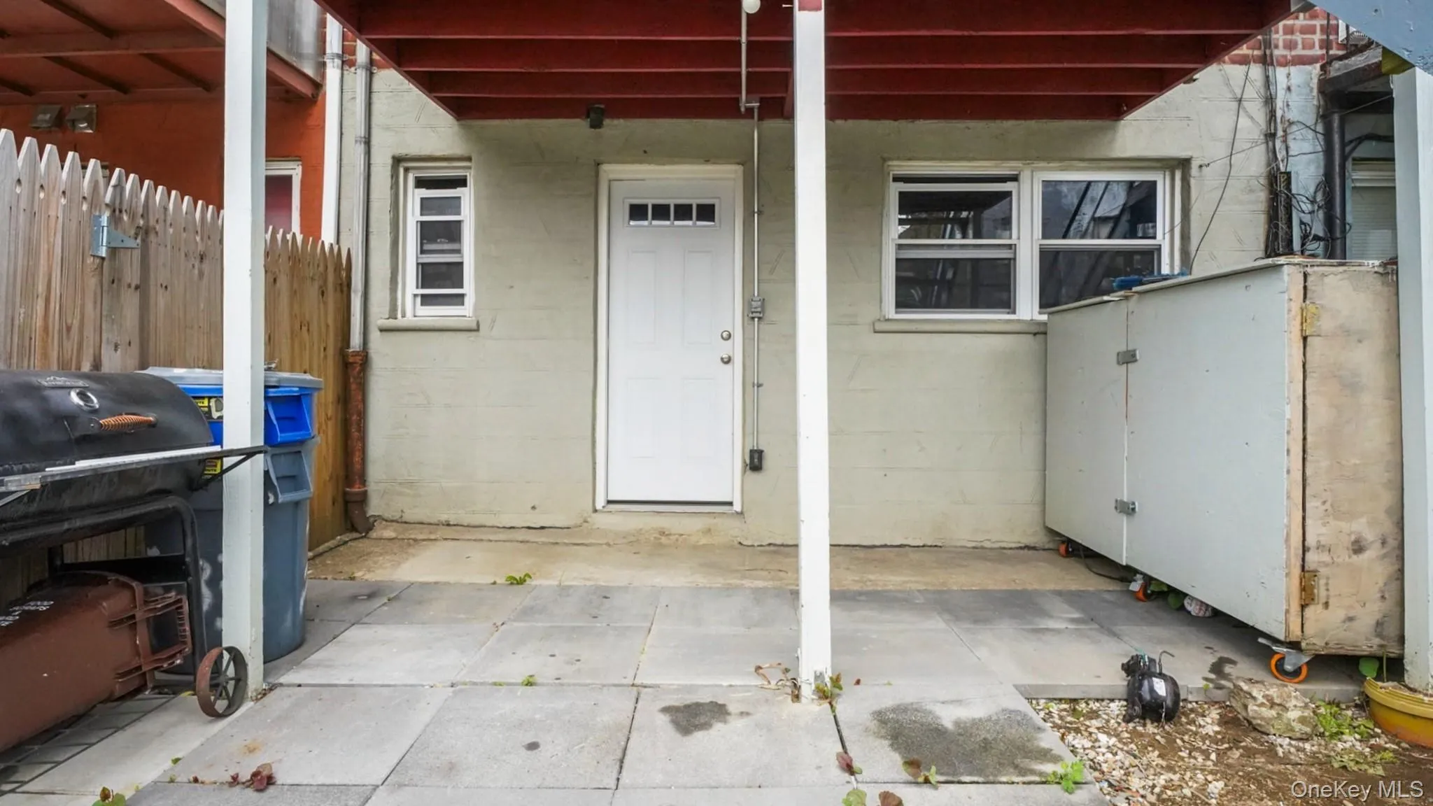 Paved outdoor patio area featuring a white door with five-pane window, two white-framed windows, and a partially visible wood fence Paved outdoor patio area featuring a white door with five-pane window, two white-framed windows, and a partially visible wood fence