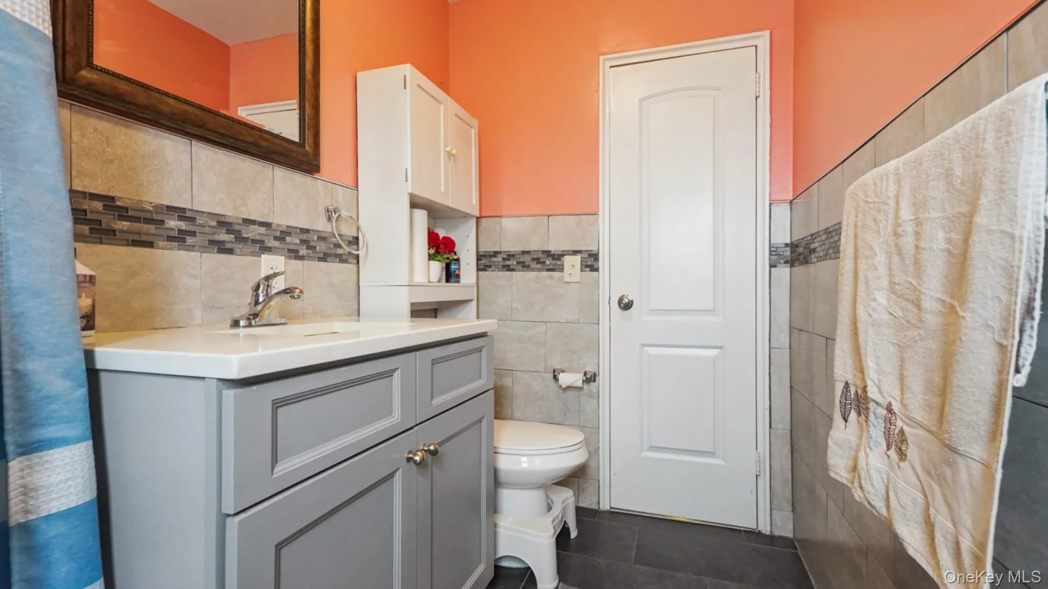 Bathroom featuring a gray vanity with a white countertop, tiled walls with a decorative mosaic accent, dark tile flooring, and a white paneled door Bathroom featuring a gray vanity with a white countertop, tiled walls with a decorative mosaic accent, dark tile flooring, and a white paneled door